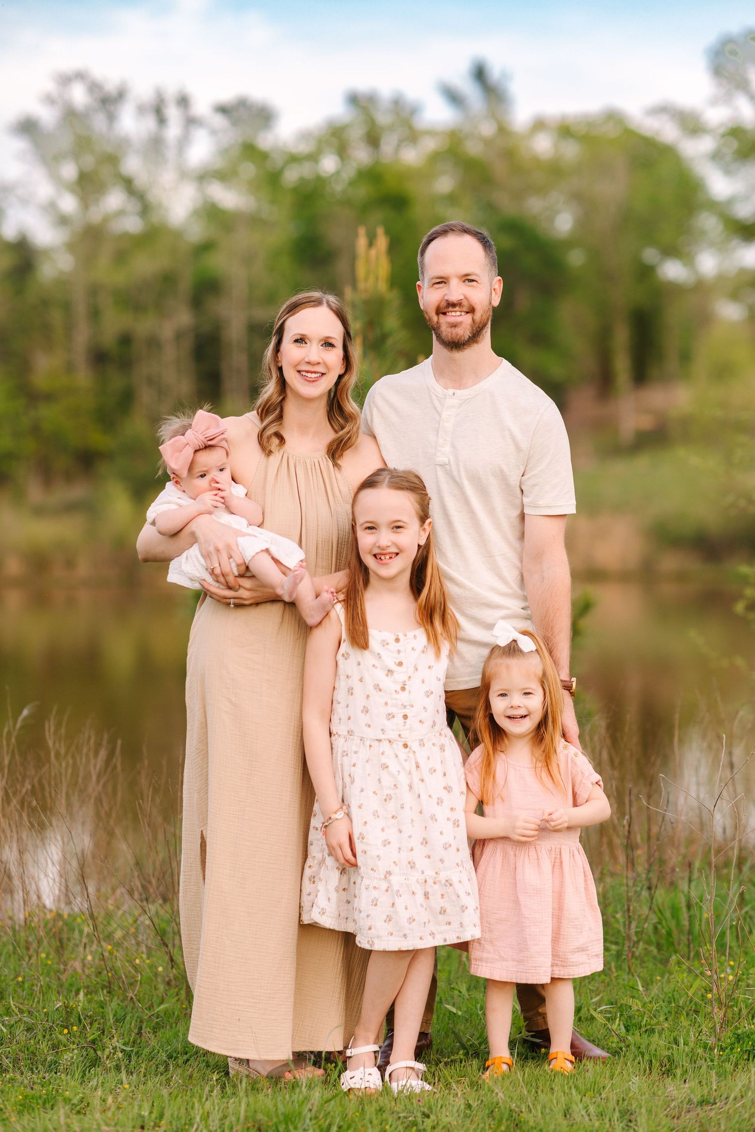 A family wearing coordinated beige and pink outfits poses together by a lake during sunset.