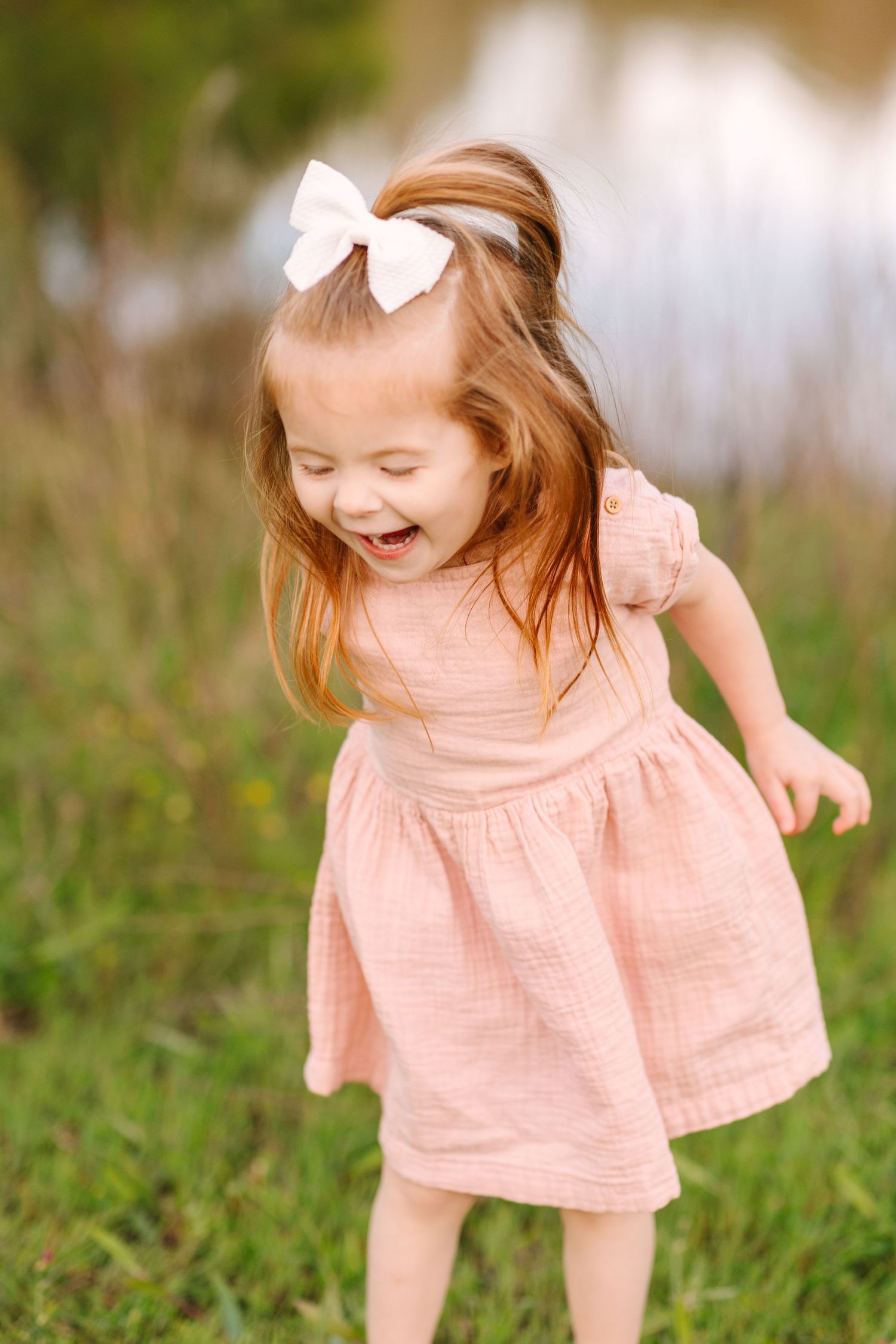 A young child in a peach dress plays joyfully in a grassy outdoor setting with a white bow accessory.