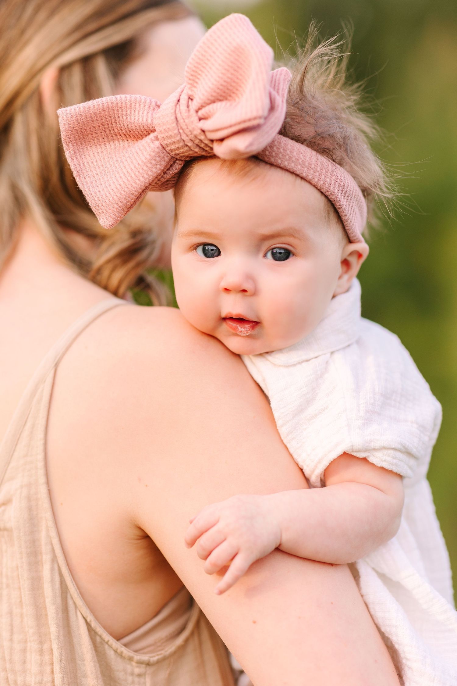 A baby in white clothing and pink bow headband being held against soft green natural background in series of photos.