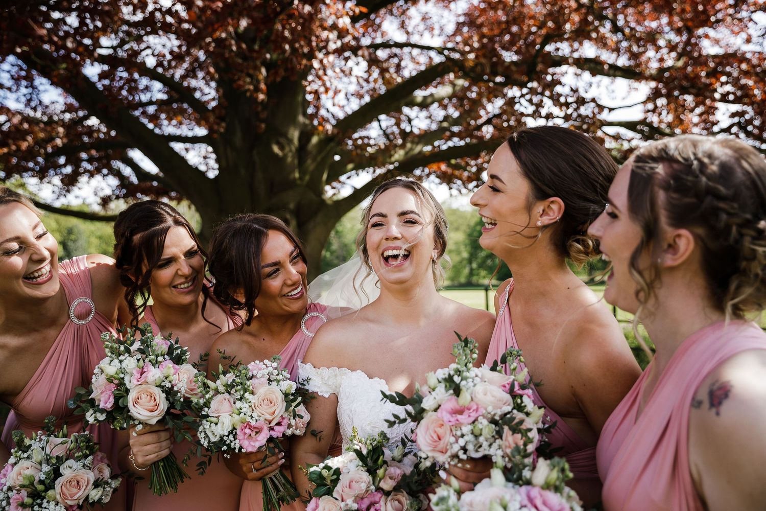 Bride and bridesmaids huddled together laughing outdoors at Kelham House, wearing pink dresses and holding floral bouquets
