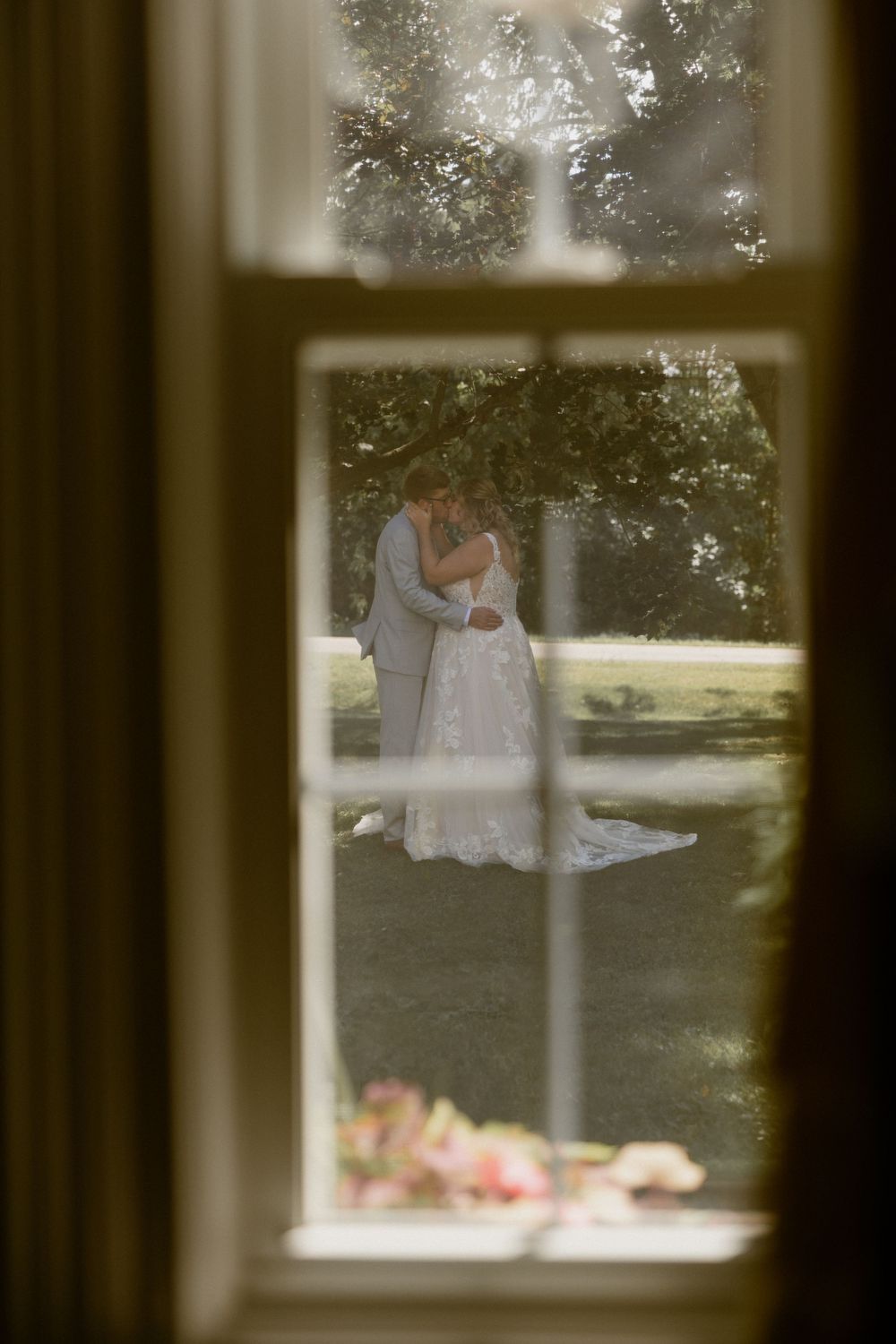 Through a window frame, a peaceful soft focus scene shows two people in white sharing an intimate outdoor moment.