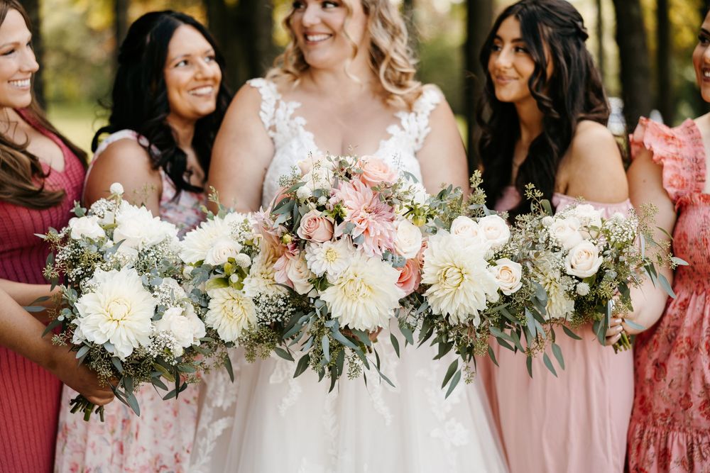 A wedding party holds beautiful bouquets of white dahlias, blush pink roses and cascading greenery in coordinating dresses.