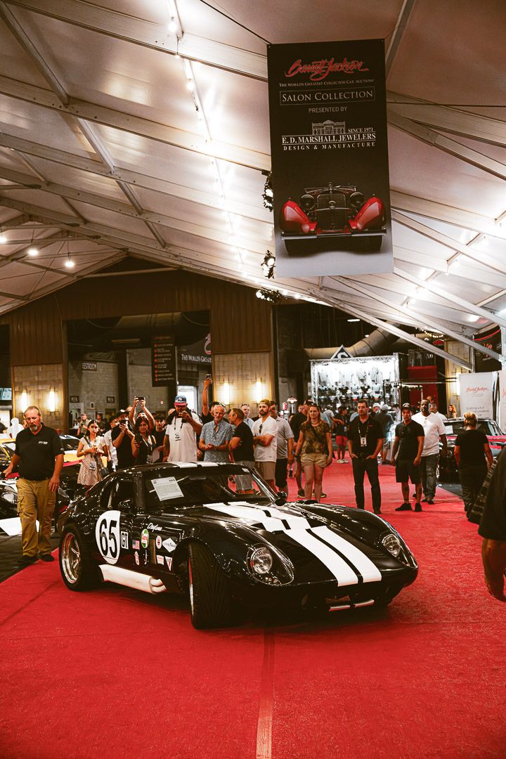Luxury vehicles showcased on red carpet in a large white exhibition tent with overhead lighting.