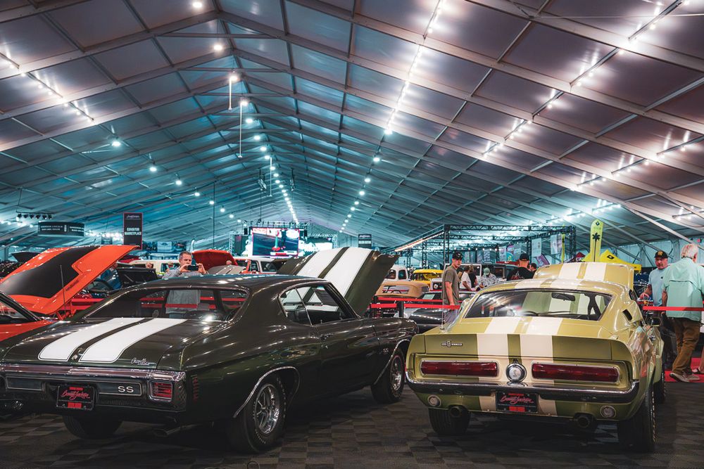 Classic muscle cars and vintage vehicles on display in a large indoor exhibition hall with bright lighting and vaulted ceiling.