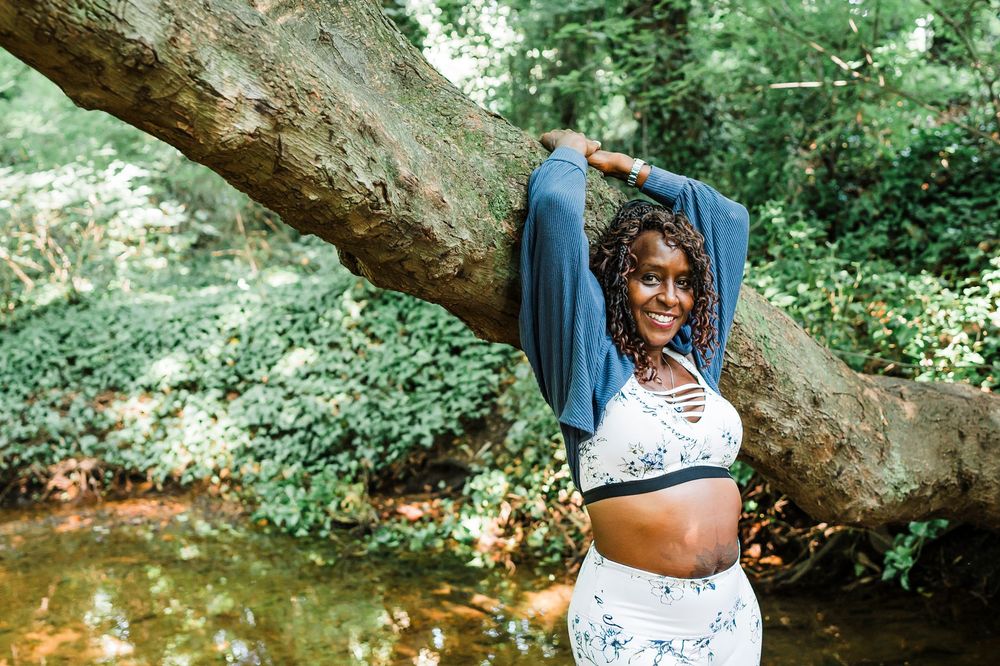 A person in white dress stands joyfully by a fallen tree trunk in a lush green forest setting.