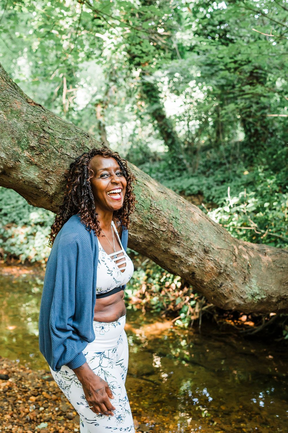 Someone laughs joyfully while sitting near a fallen tree branch by a stream on a sunny summer day.