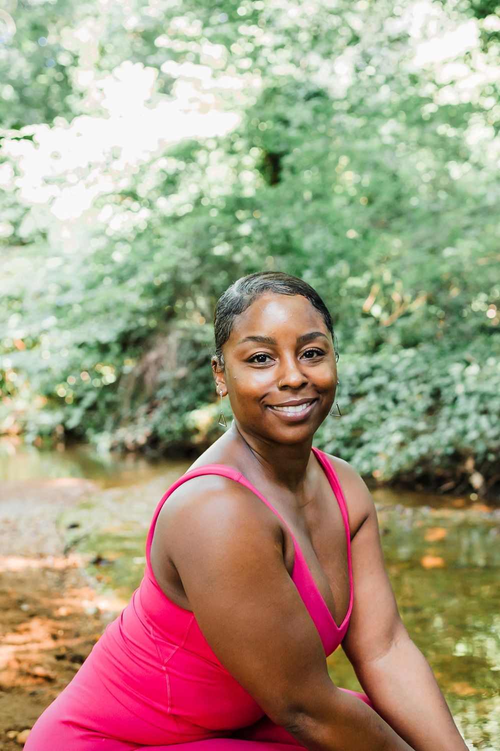 A person in a bright pink top poses outdoors near a stream surrounded by lush green foliage and water.