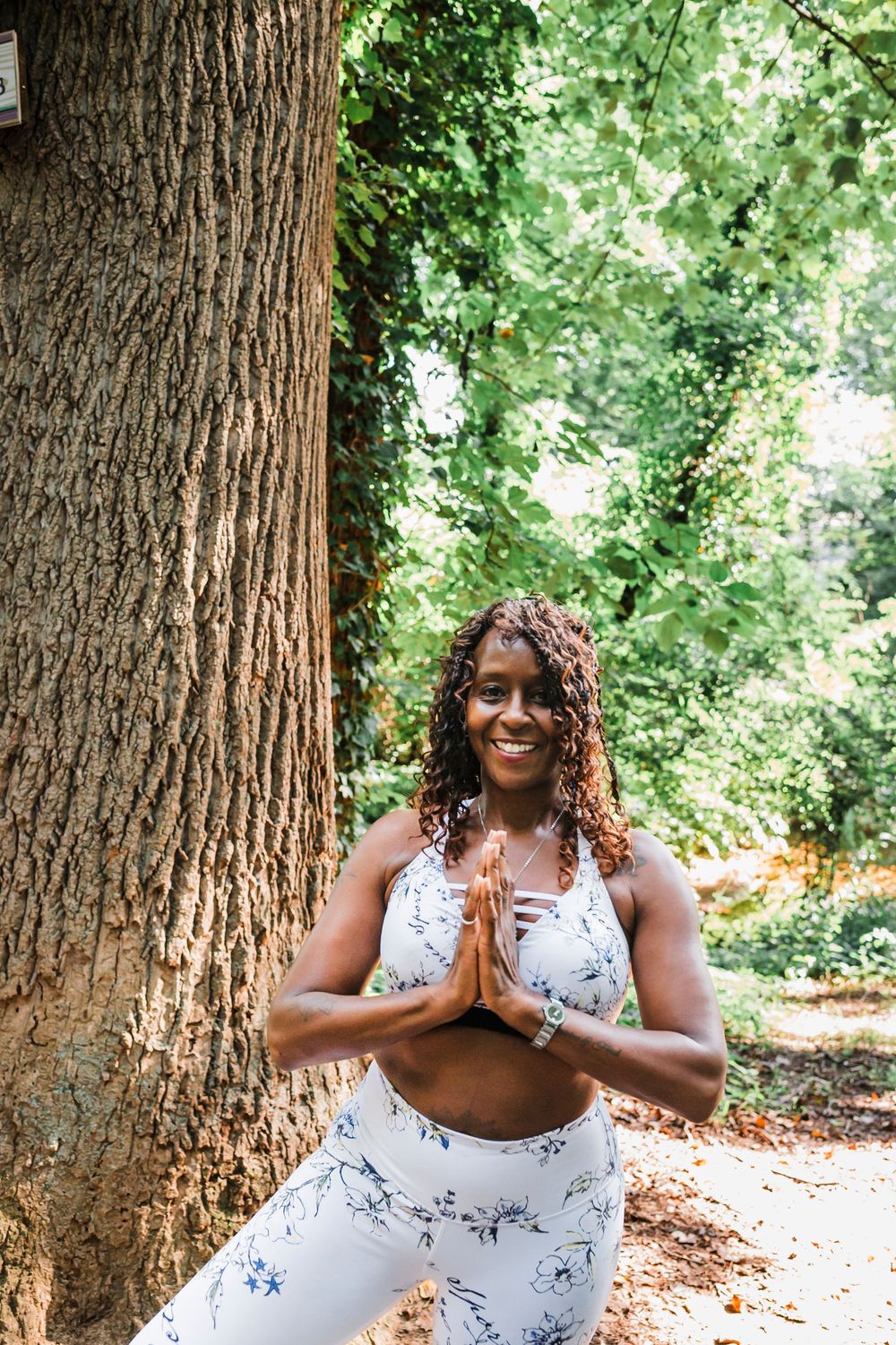 A person in a white floral dress practices peaceful meditation poses against large tree trunks in a lush forest setting.
