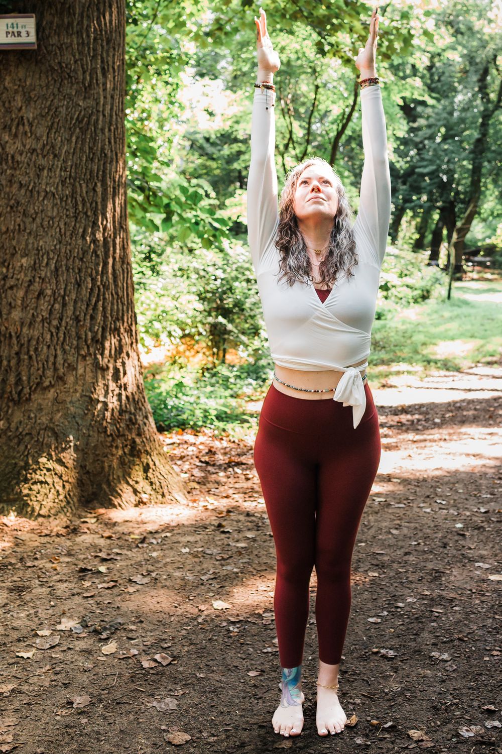 Person in maroon leggings and white crop top performing stretching exercises in a wooded outdoor setting.