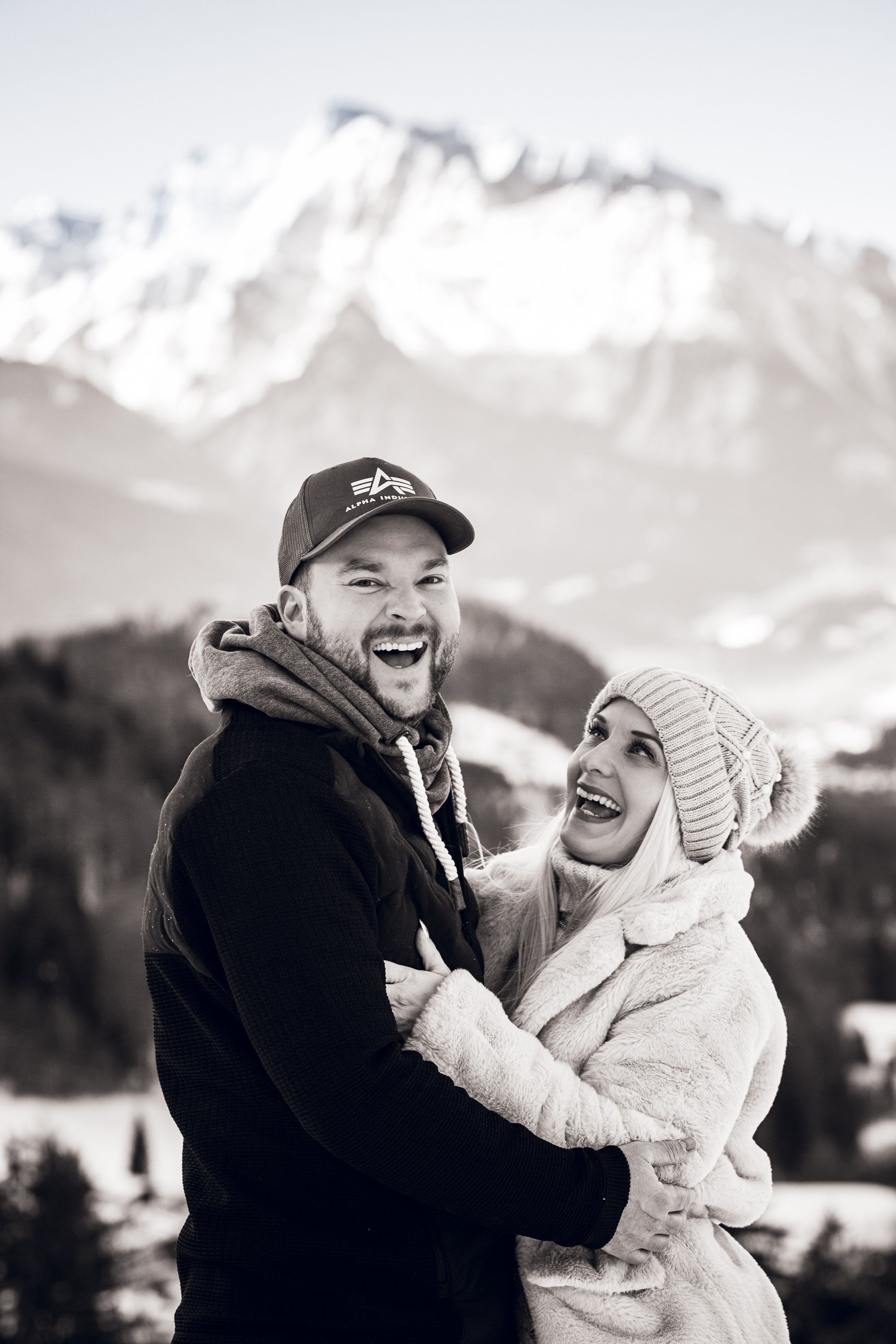 A joyful couple embraces and laughs together while standing in front of snow-capped mountain peaks on a winter day.