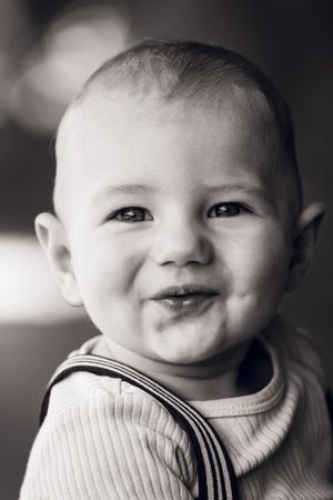 Black and white portrait of a smiling baby with chubby cheeks wearing suspenders.