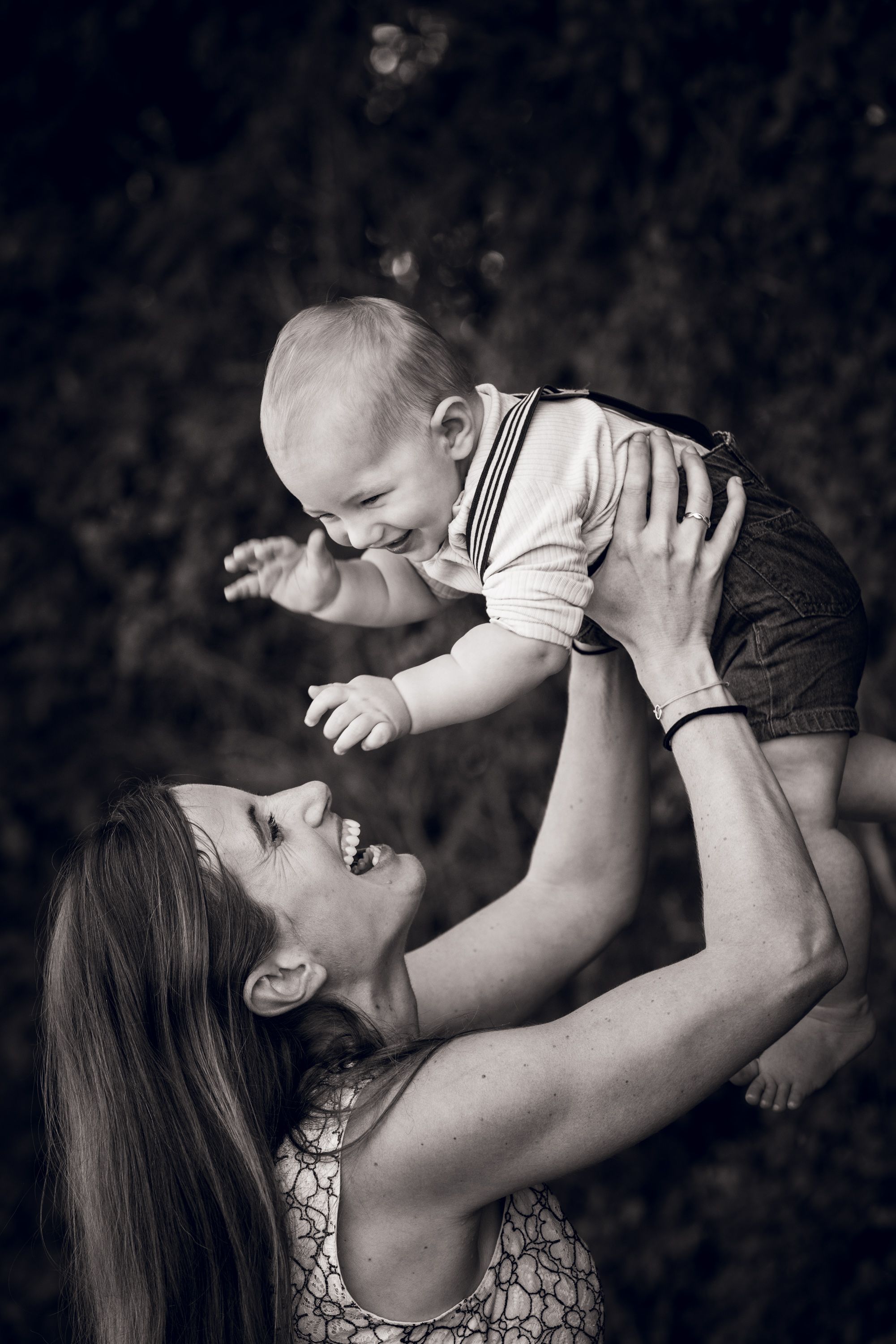Black and white photo of a joyful mother lifting her laughing baby outdoors, sharing a beautiful bonding moment.