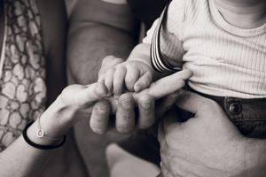 Black and white photo of a baby holding an adults finger, with two adults hands gently supporting the tender moment.