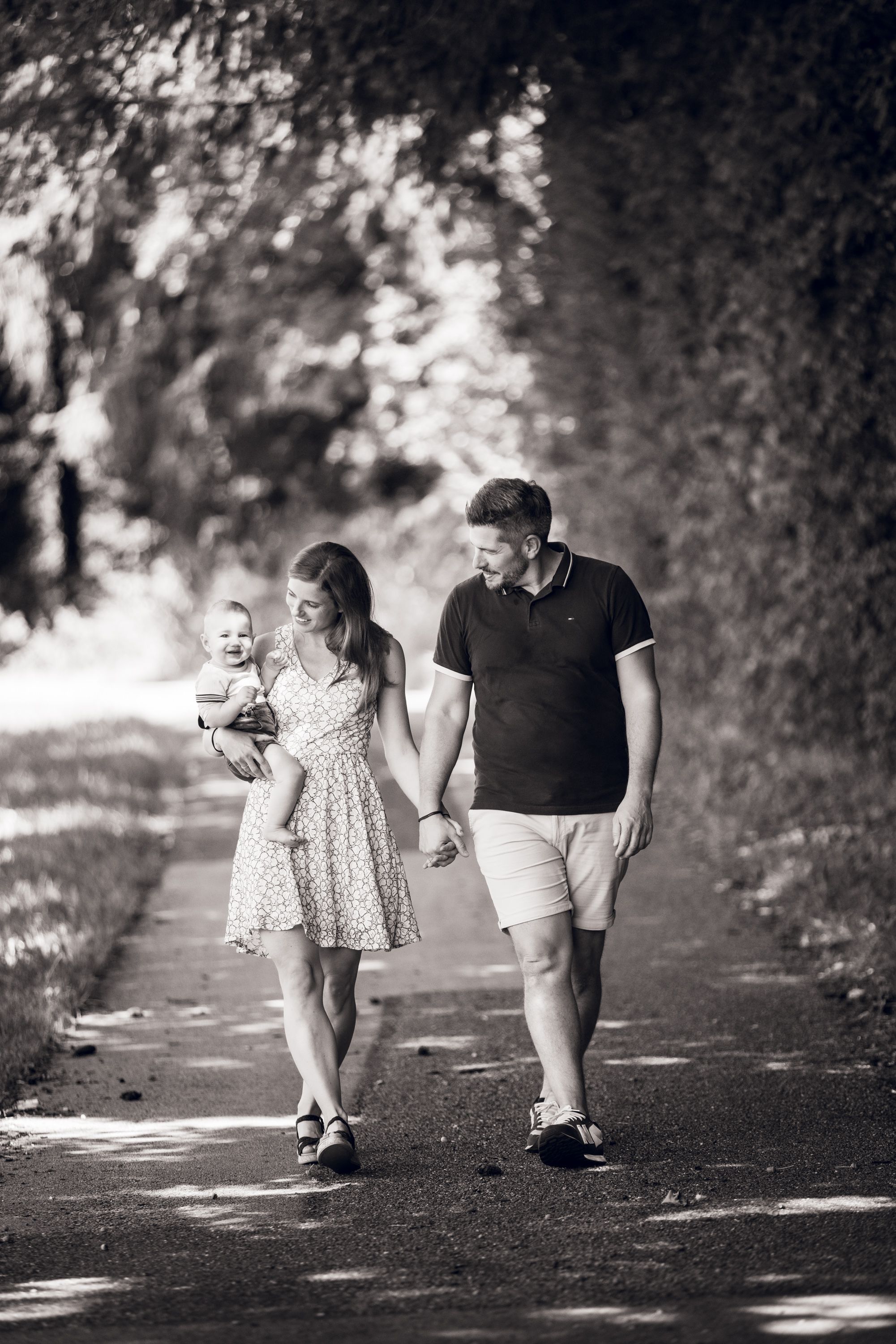 Black and white photo of a young family walking on a tree-lined path, mother holding baby while holding fathers hand.