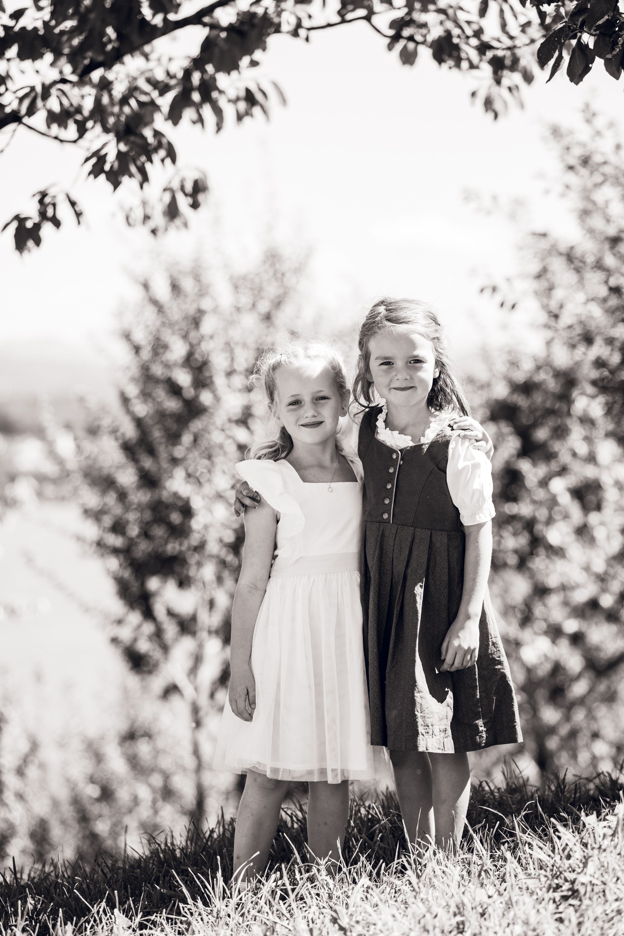 Two young girls in dresses pose together outdoors in a black and white portrait photo.