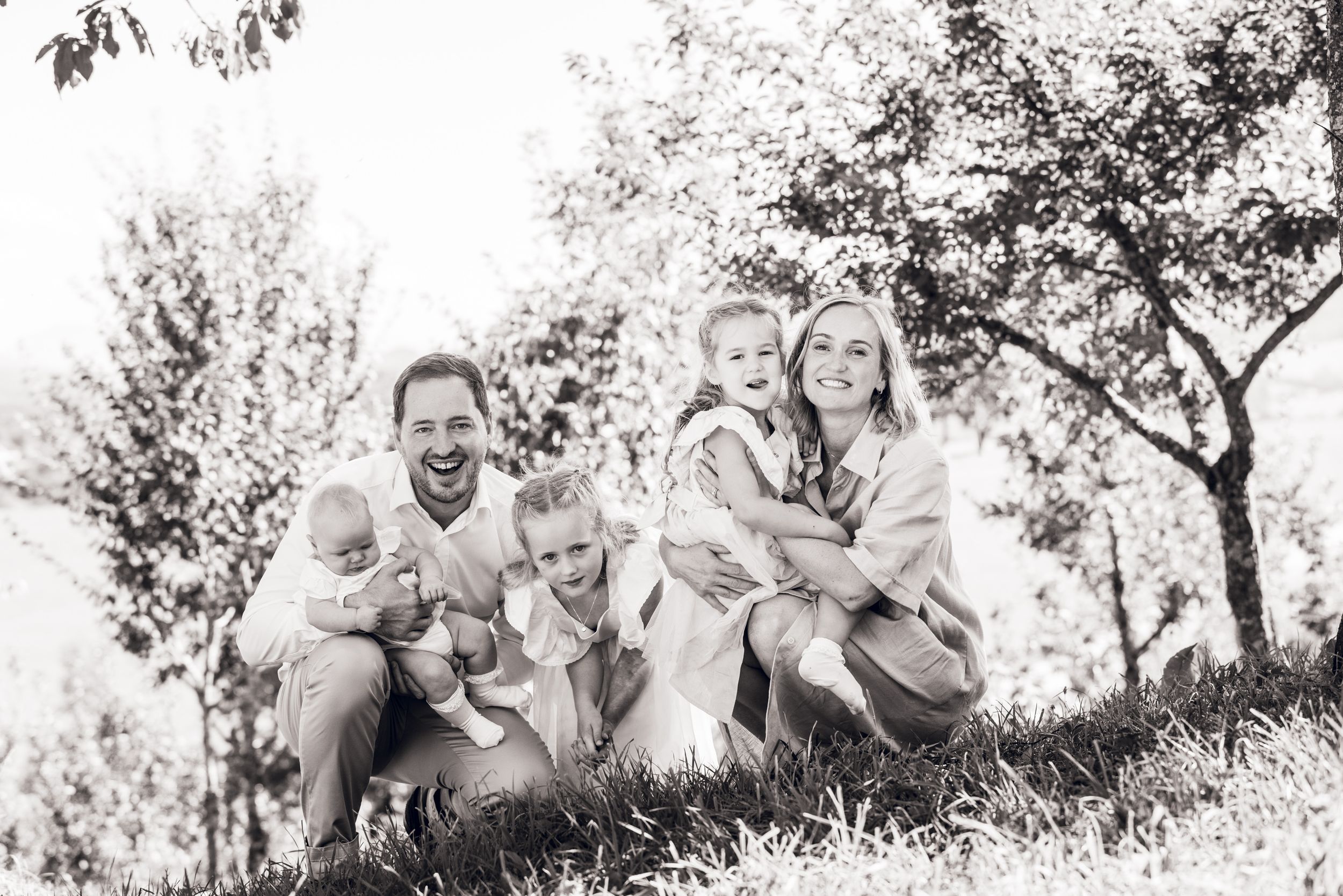 Black and white family photo of parents with three young children smiling outdoors among trees on a grassy hill.