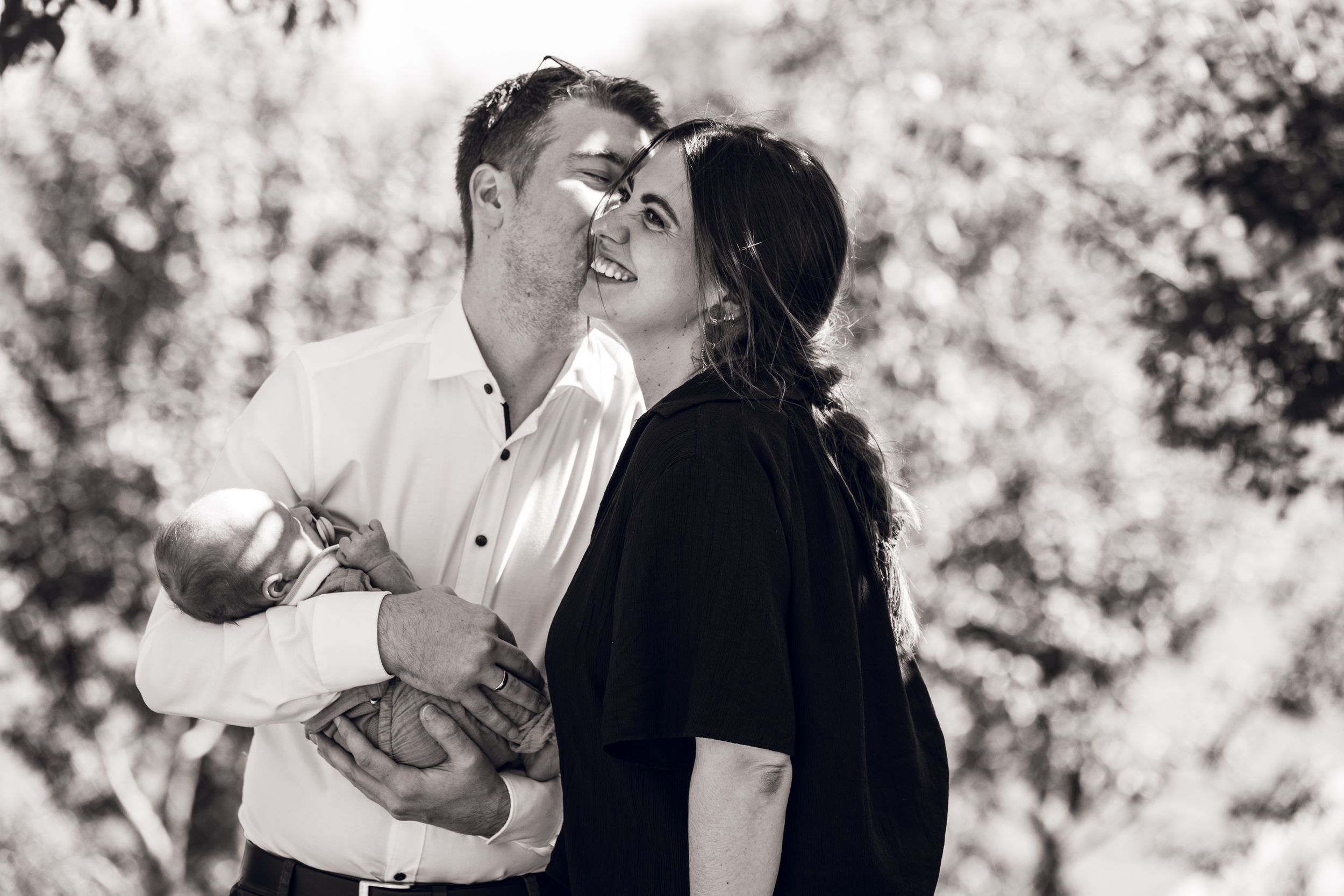 Black and white photo of a smiling couple outdoors, man holding a baby, woman in black top leaning close.