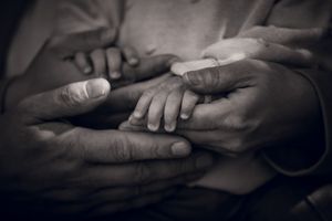 Black and white photo of multiple hands cradling a tiny newborn baby hand, symbolizing family love and new life.