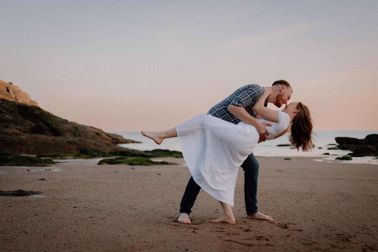 A couple in white shares an intimate dance movement together on a beach during sunset.