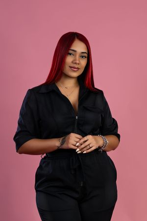 Woman with red hair wearing a black jumpsuit poses confidently against a pink background.