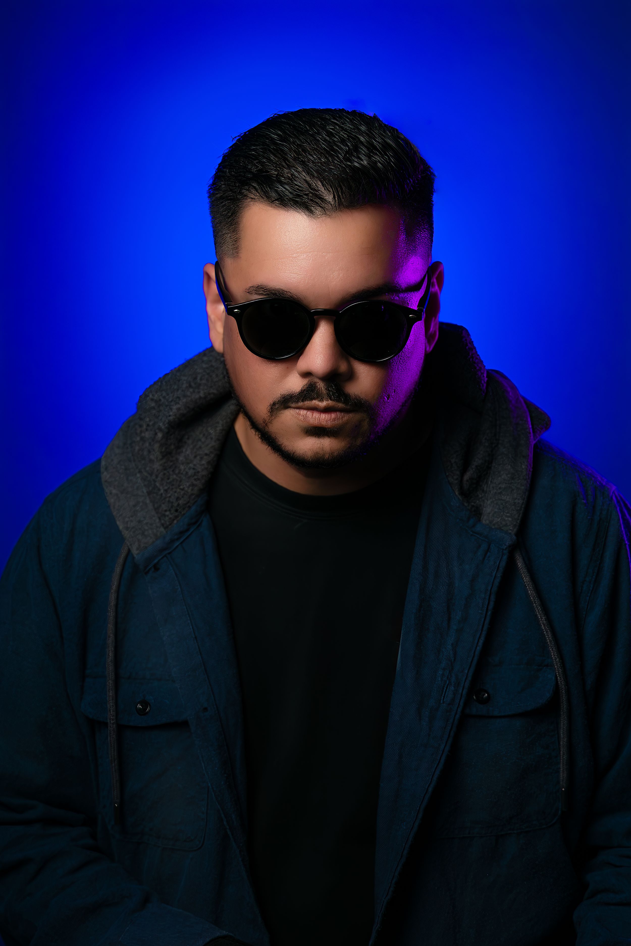 Mysterious man in dark jacket and black sunglasses poses against vibrant blue background with purple lighting.