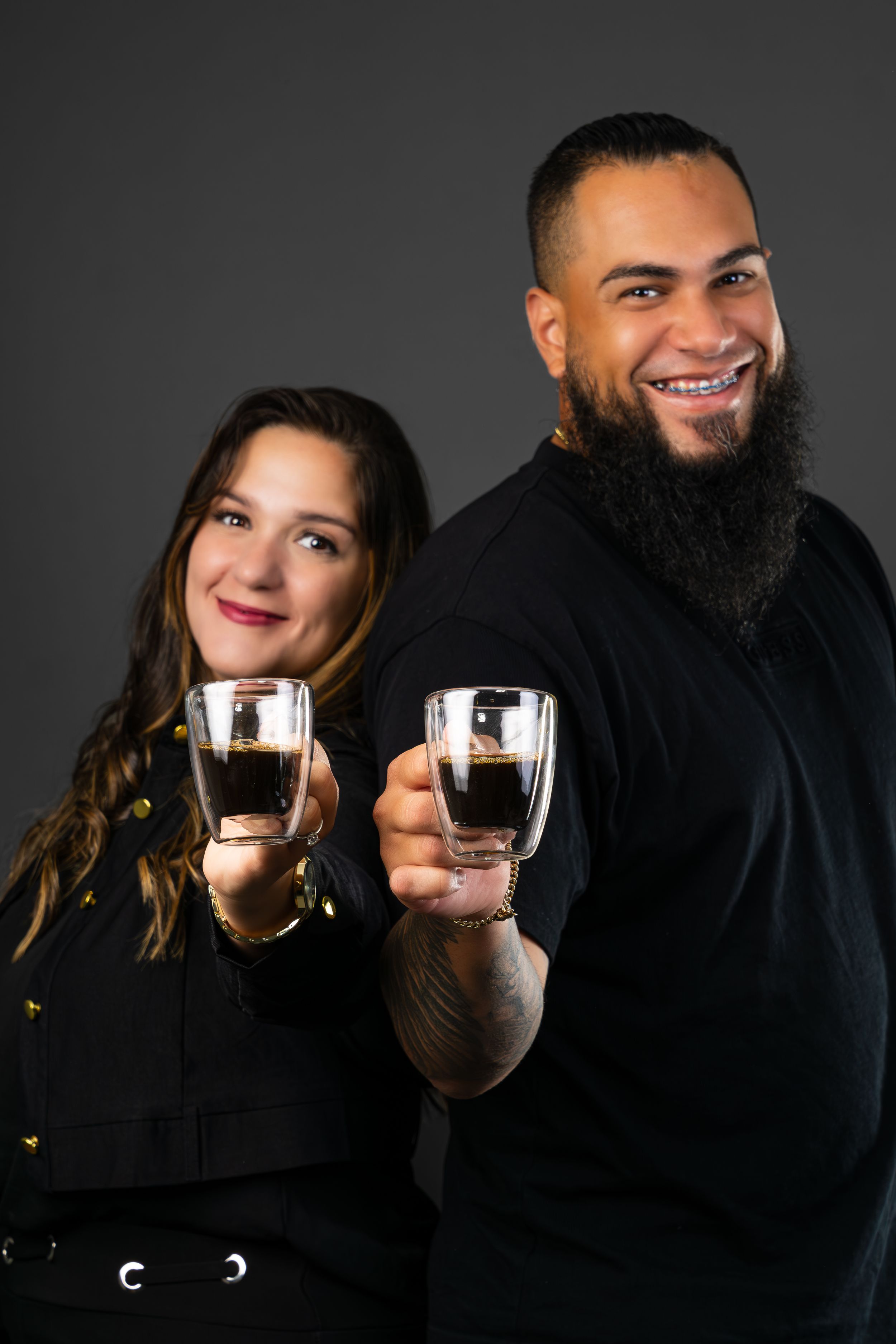 Smiling couple in black outfits toasting with espresso shot glasses in a studio portrait.