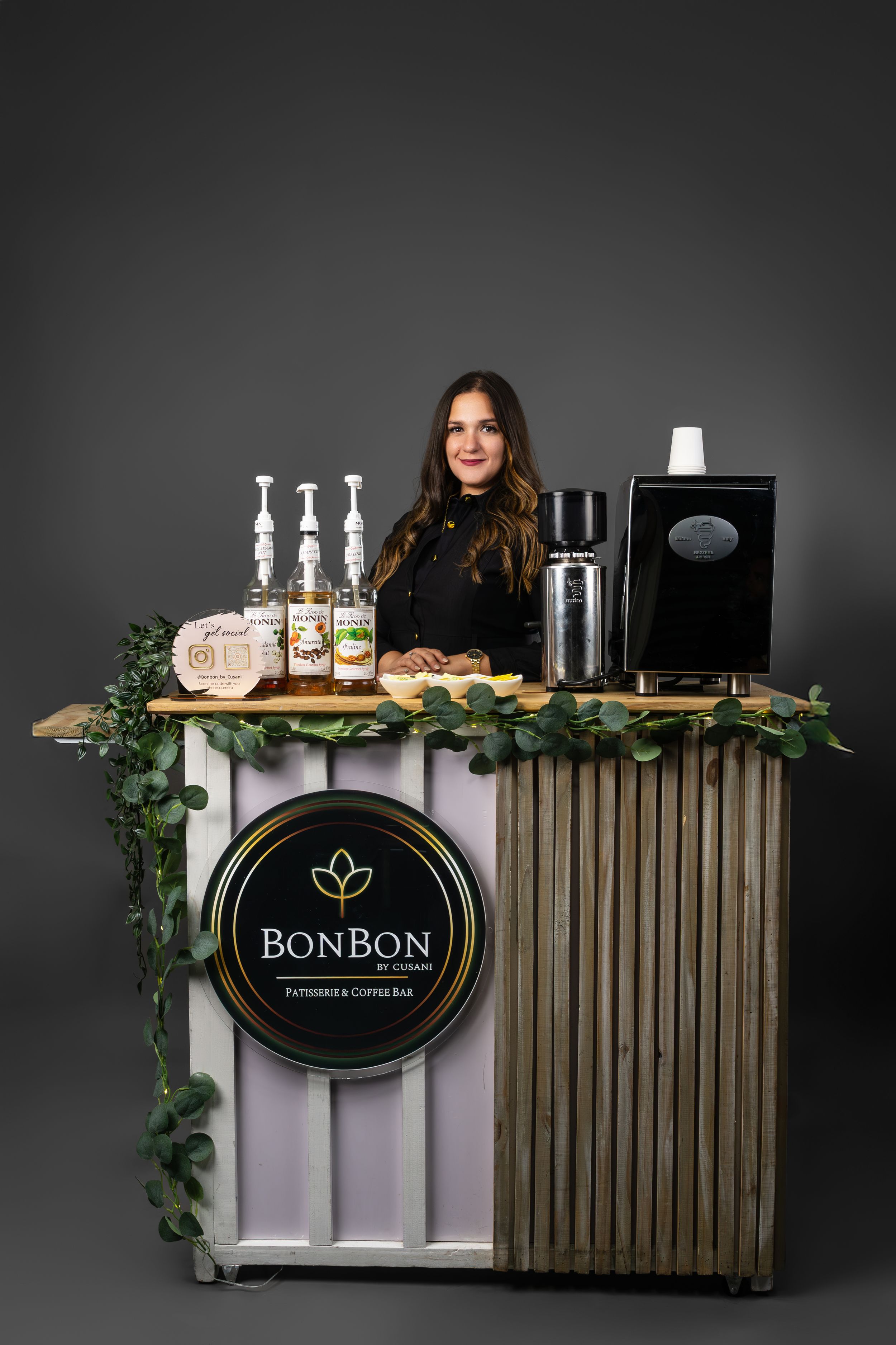 Woman stands behind BonBon Patisserie and Coffee Bar cart with coffee equipment and green garland decor.