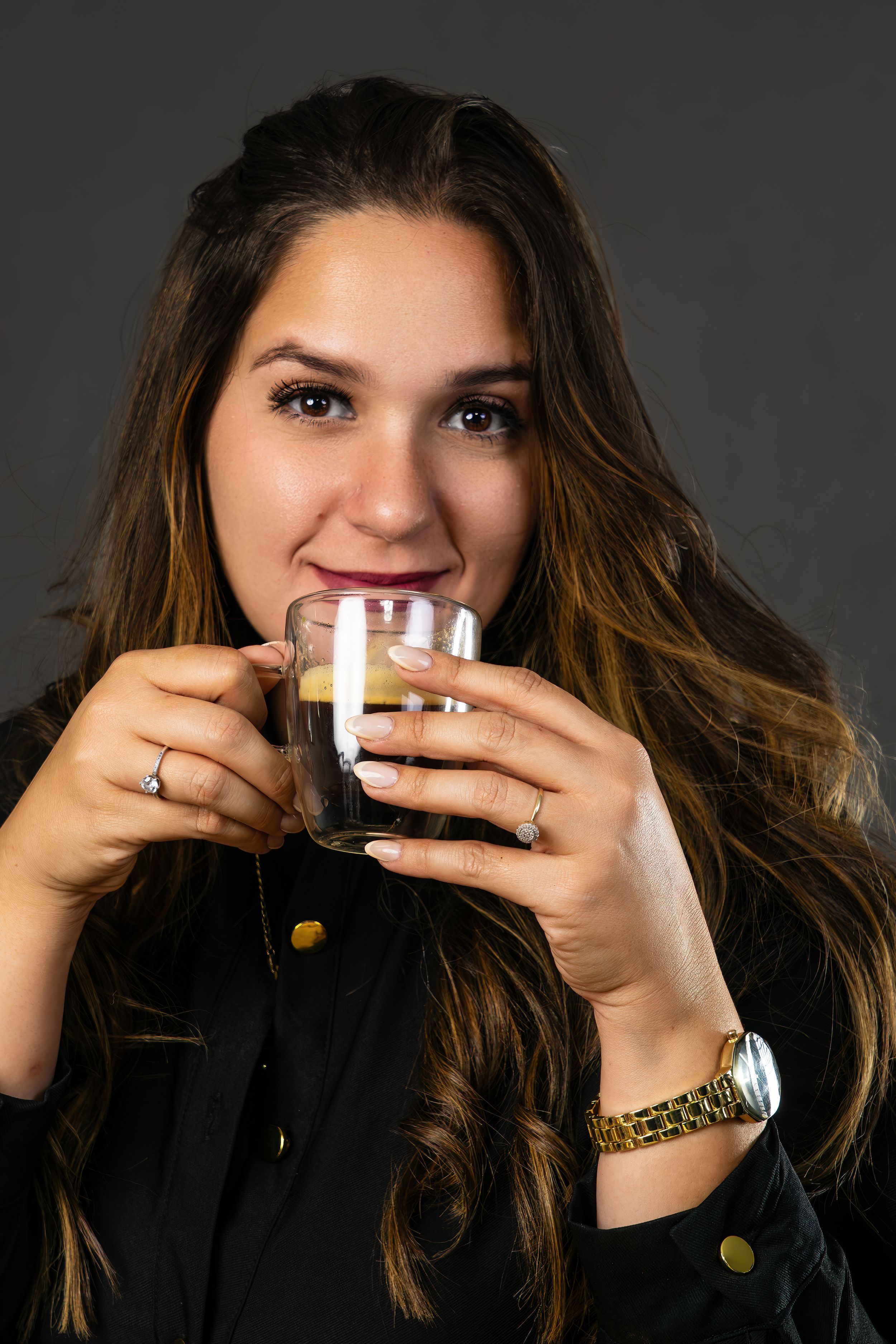 Young woman with long brown hair smiles while sipping espresso from a clear double-wall glass.