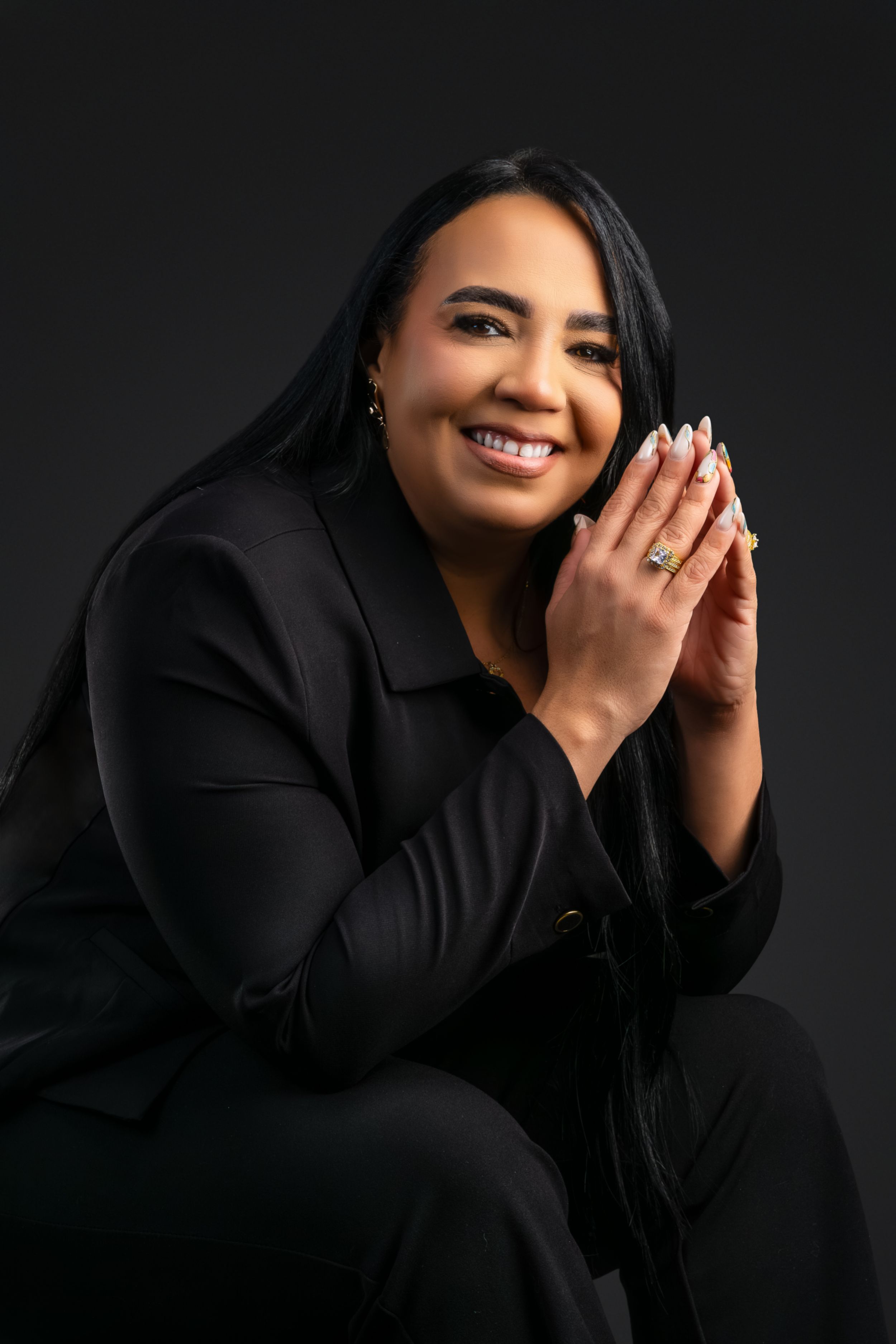 Smiling woman in black suit poses with hands together in professional studio portrait.