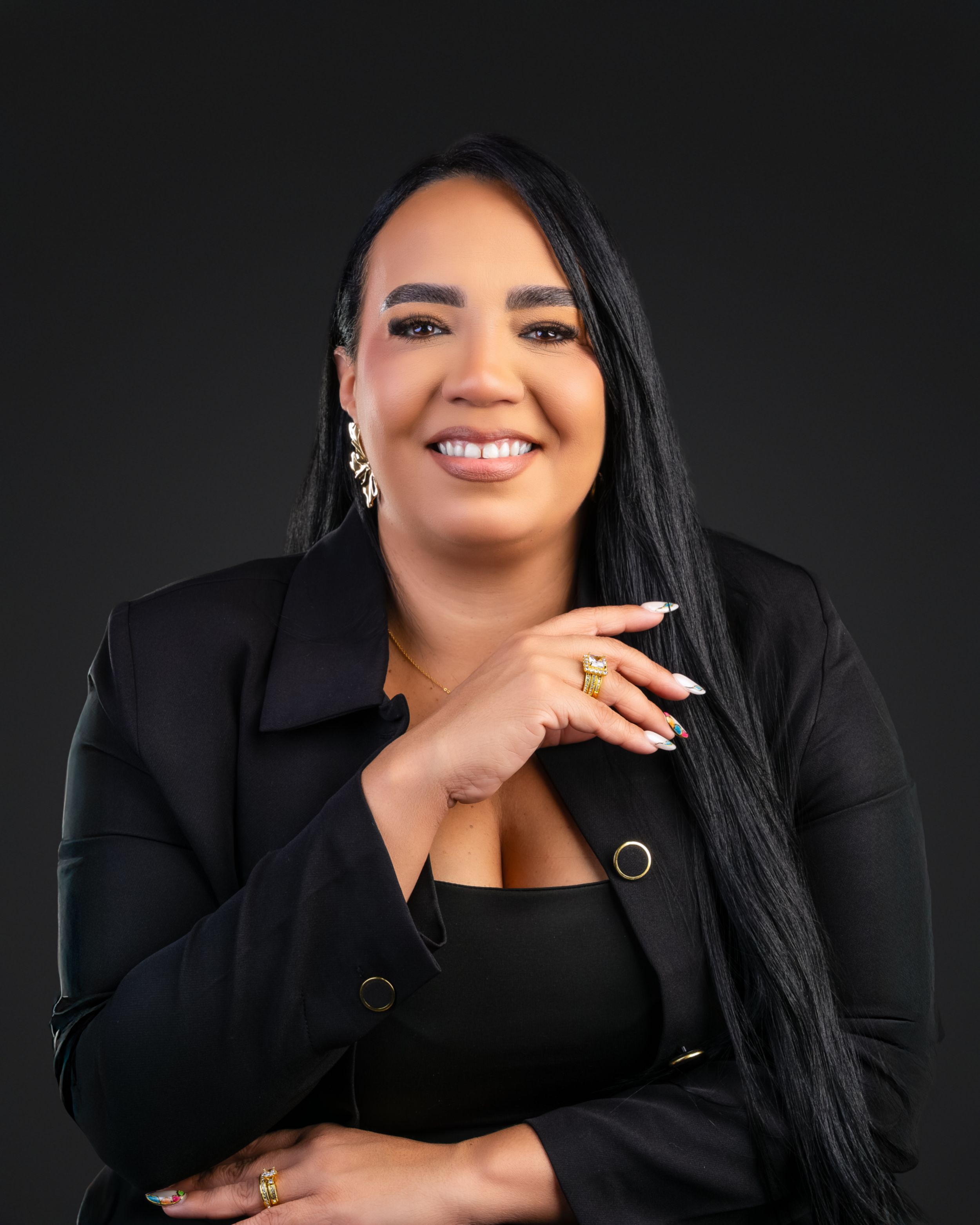 Confident woman in black blazer smiles warmly in professional headshot against dark background.