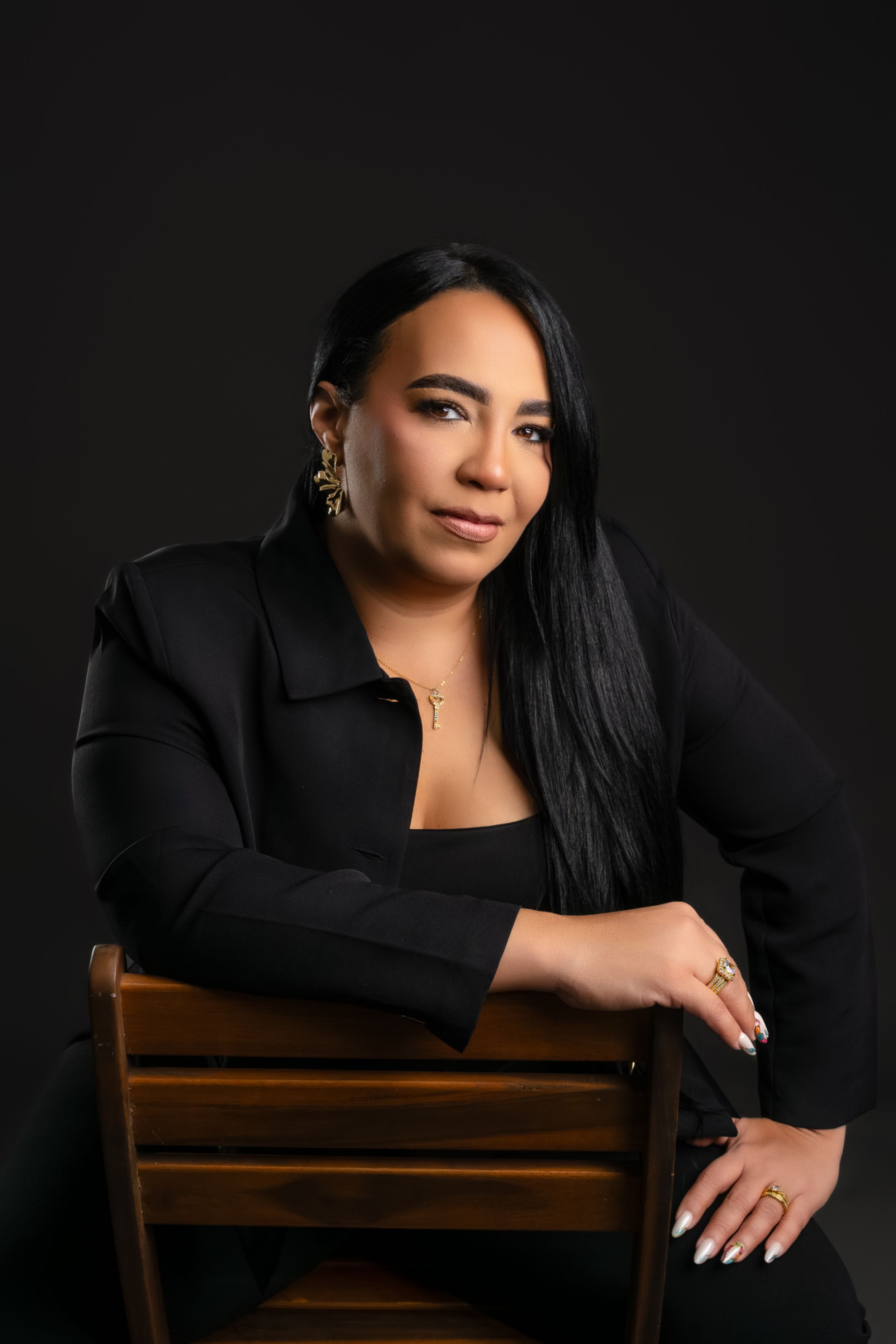 Professional woman in black blazer poses confidently on wooden chair against dark background.