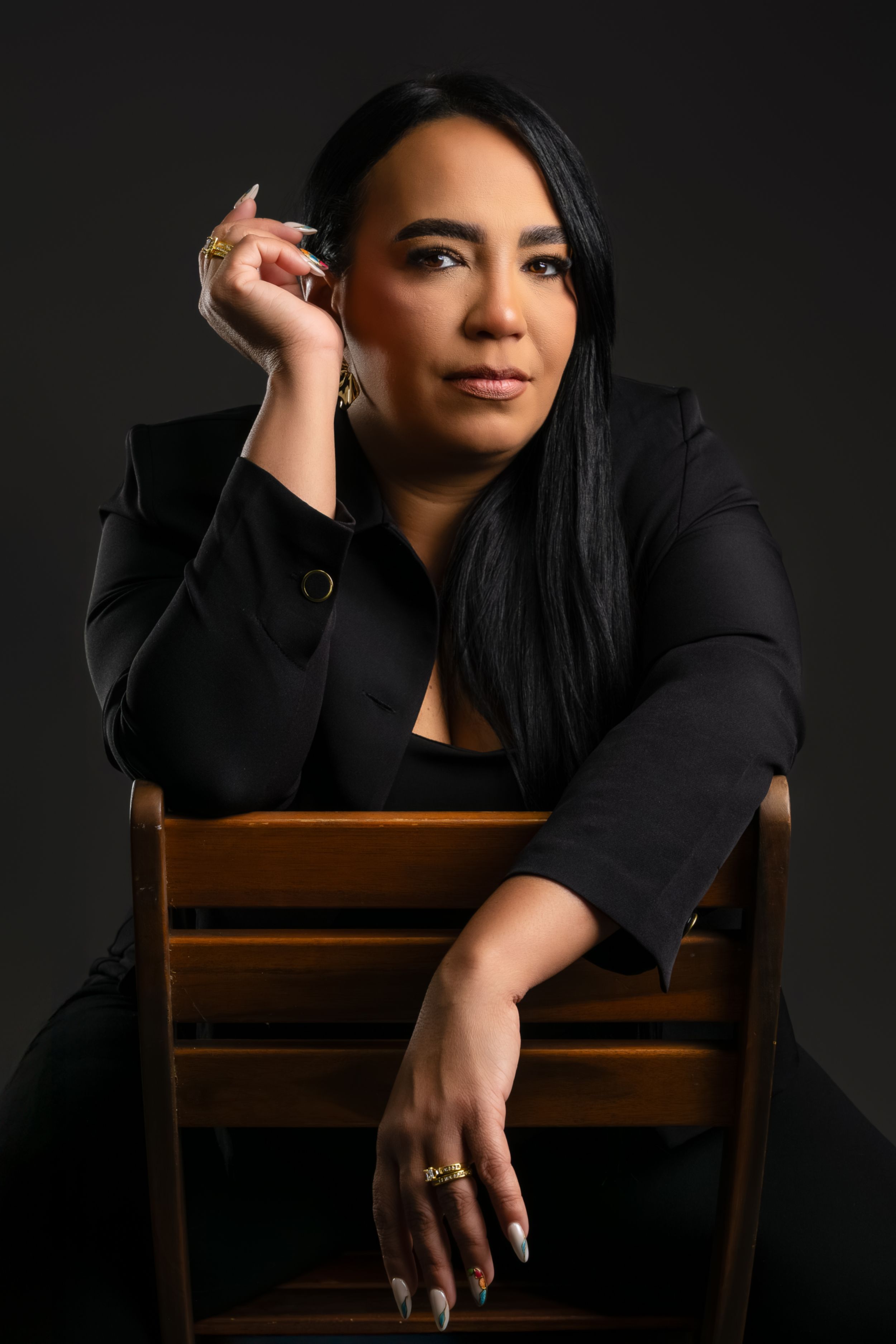 Elegant woman in black blazer sits thoughtfully on wooden chair in studio portrait.