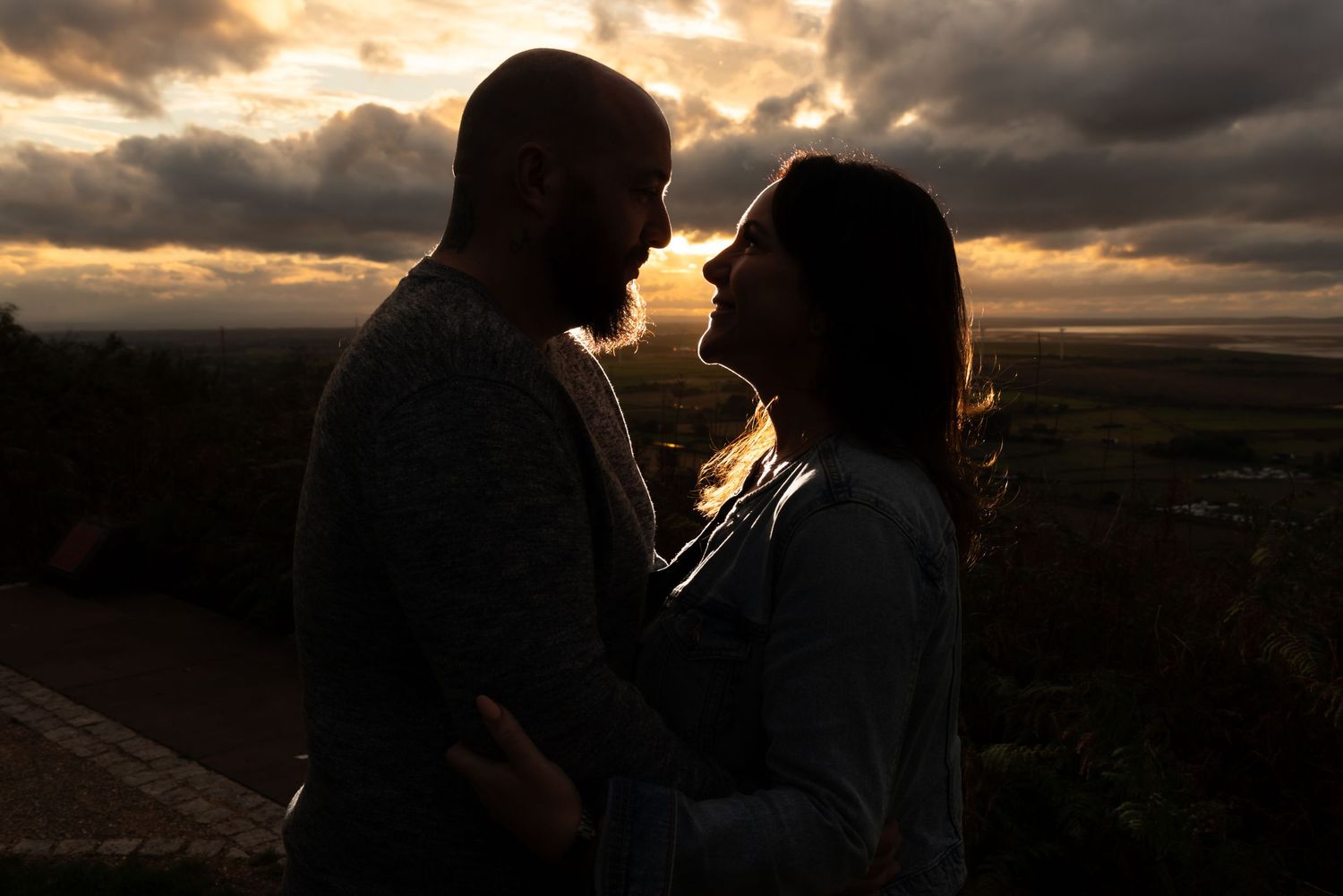 Silhouetted figures share an intimate moment against a dramatic sunset sky with dark clouds, pre wedding photography by Michael Pardoe