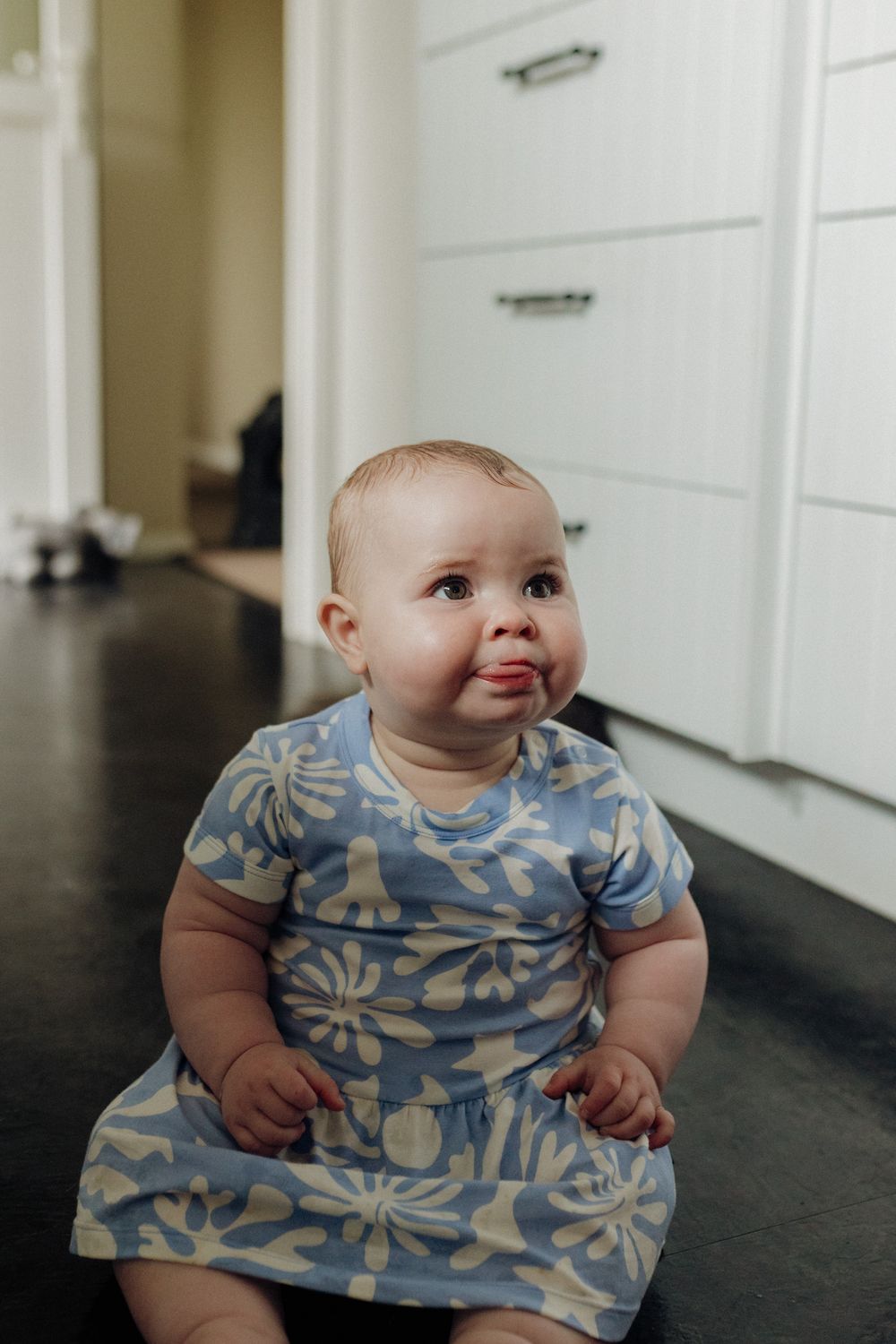 A baby in a blue floral dress sitting on a dark floor, shown in sequential photos expressing different emotions.