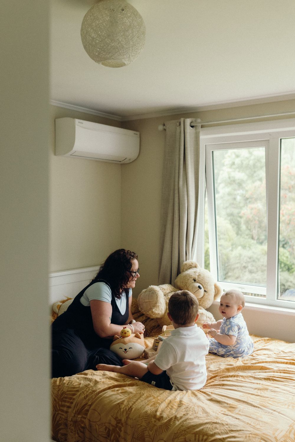 People relaxing with stuffed animals on a large bed near a bright window in a minimalist bedroom setting.