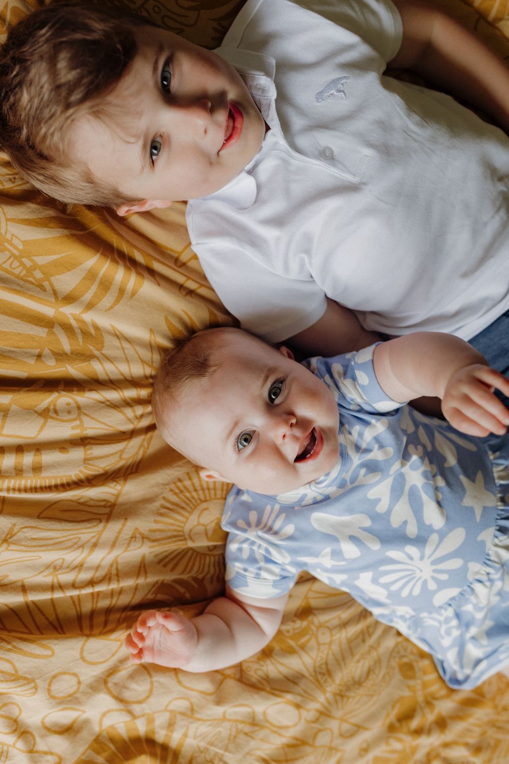 Young child lying on yellow patterned bedding wearing blue and white floral clothing while smiling.