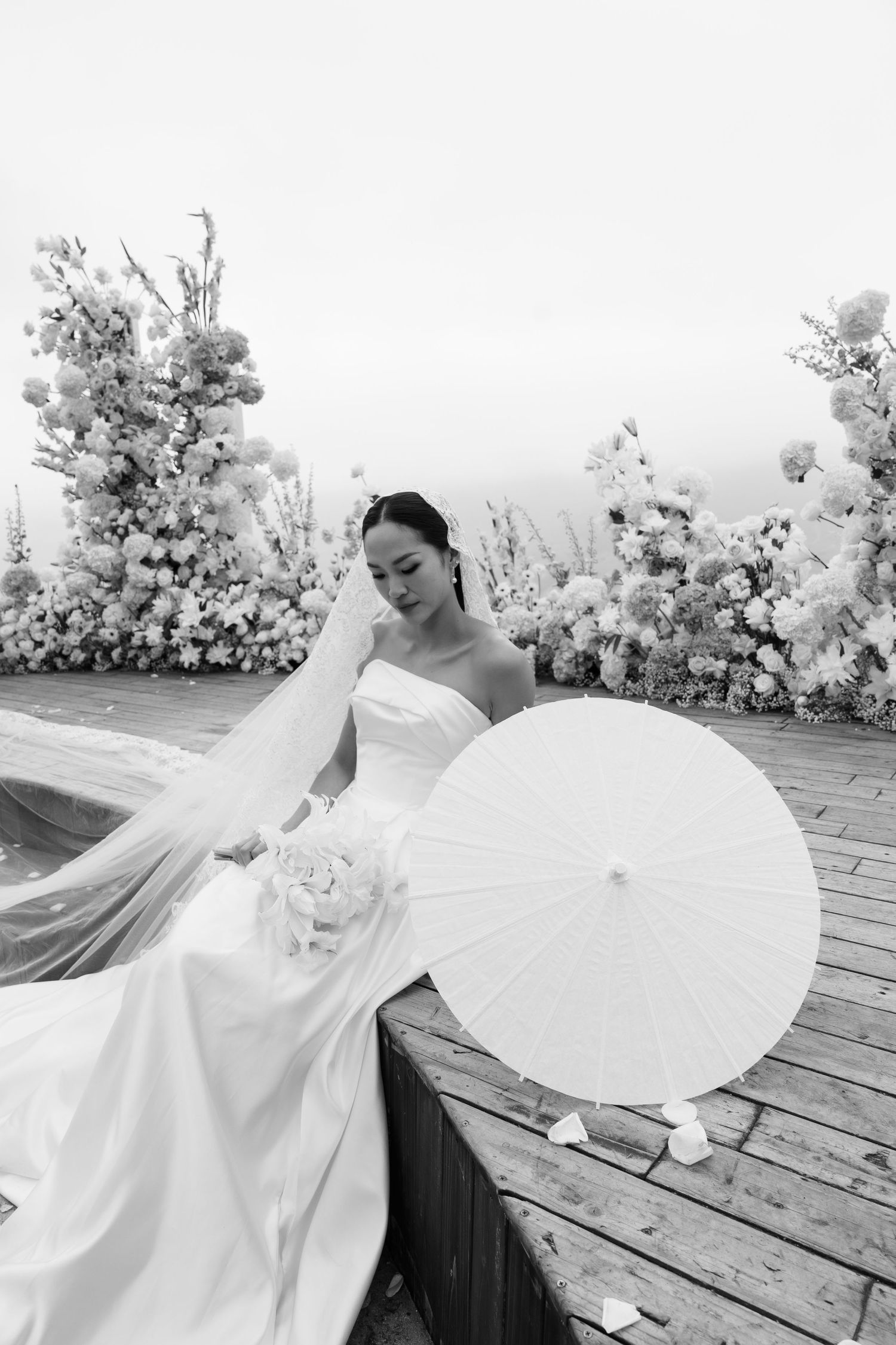 Elegant bride in white strapless gown holding parasol and bouquet seated among lush floral arrangements.