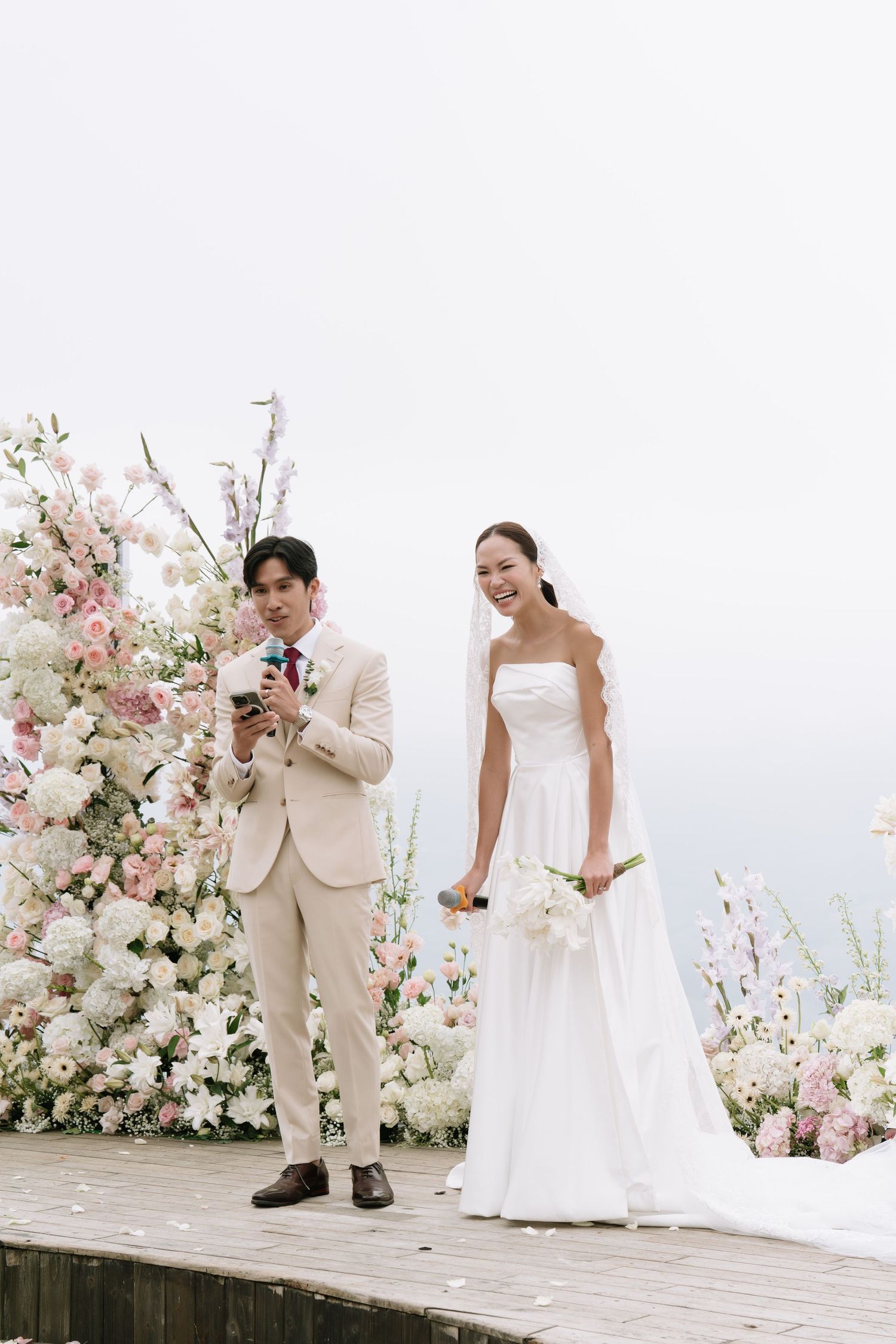 Groom in beige suit reading vows beside smiling bride holding bouquet at an outdoor floral wedding ceremony.
