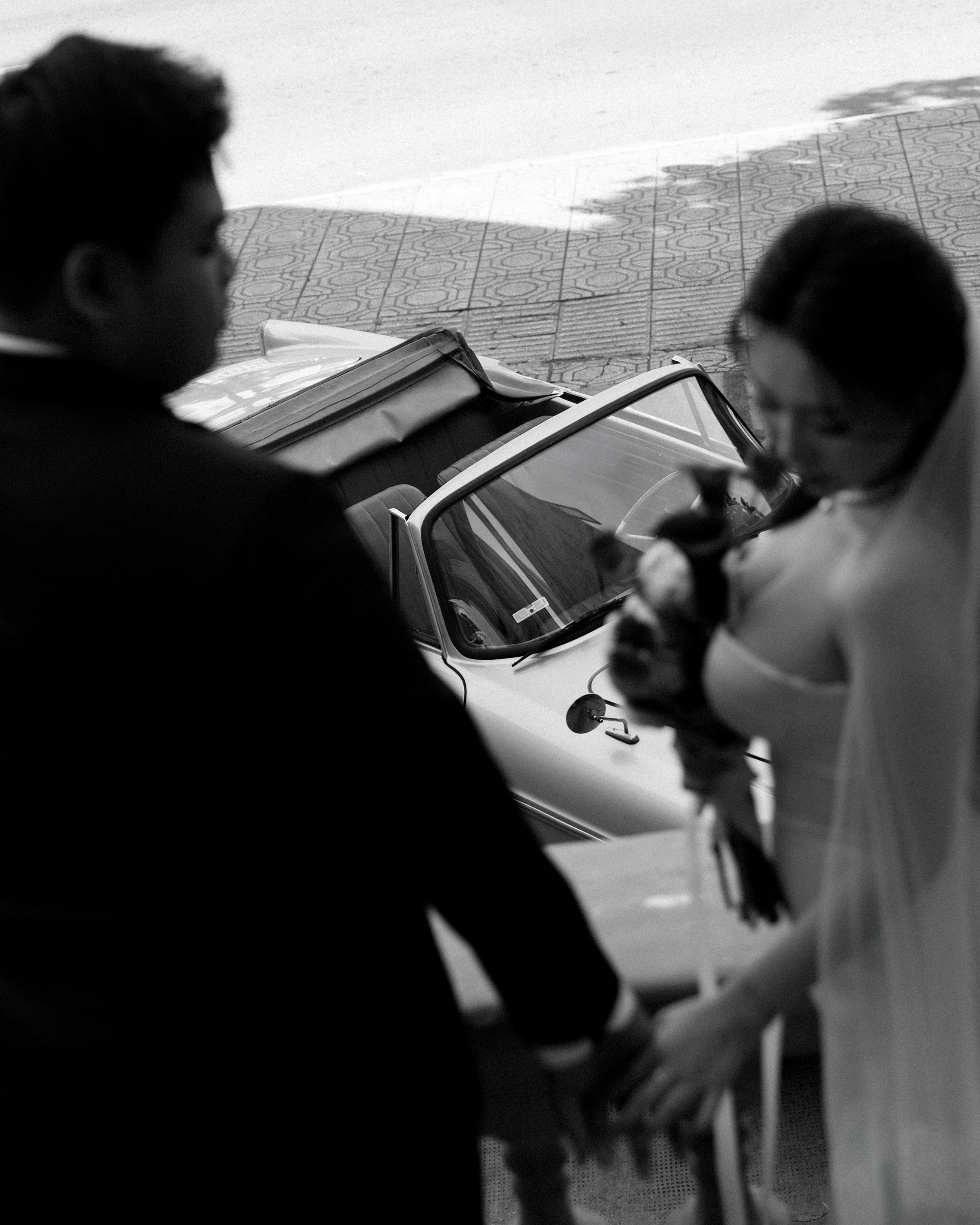 Black and white photo of bride and groom walking to a classic car on their wedding day.