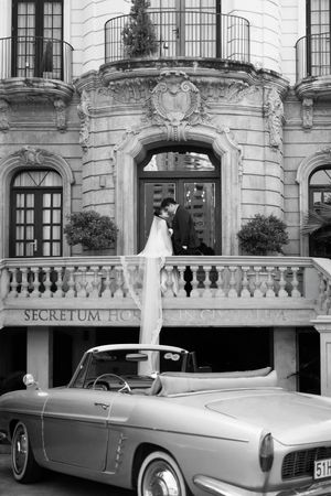 Black and white wedding photo of couple on balcony above vintage convertible at Secretum Hotel.