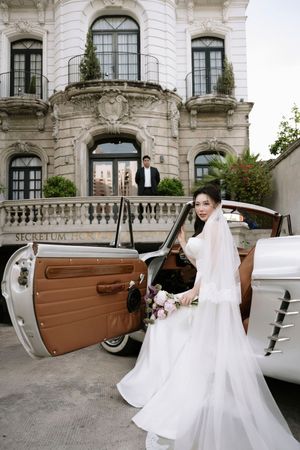 Elegant bride in white gown holds bouquet beside vintage white car at Secretum Hotel entrance.
