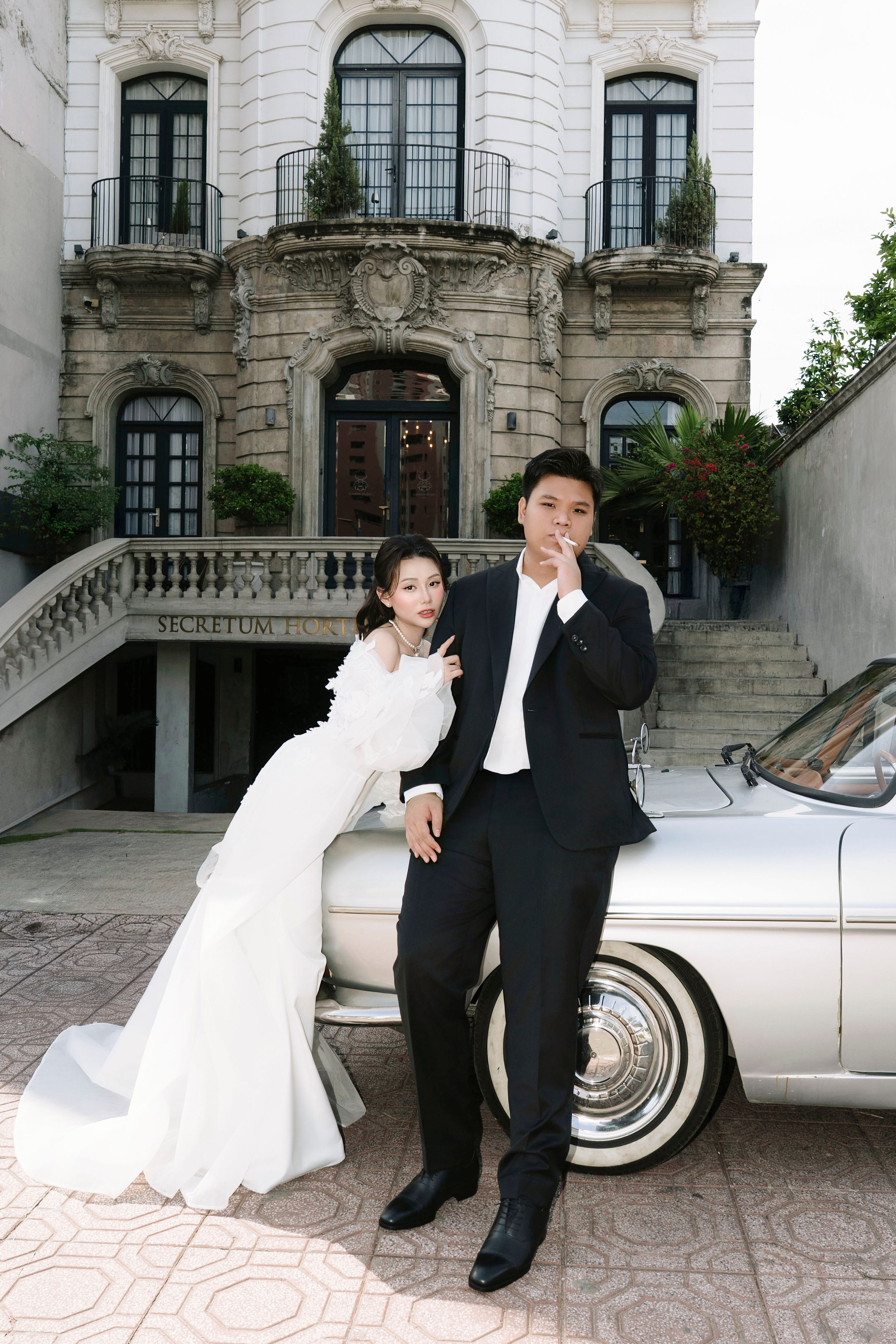 Bride in mermaid gown and groom in tuxedo pose beside vintage white car at Secretum Hotel.