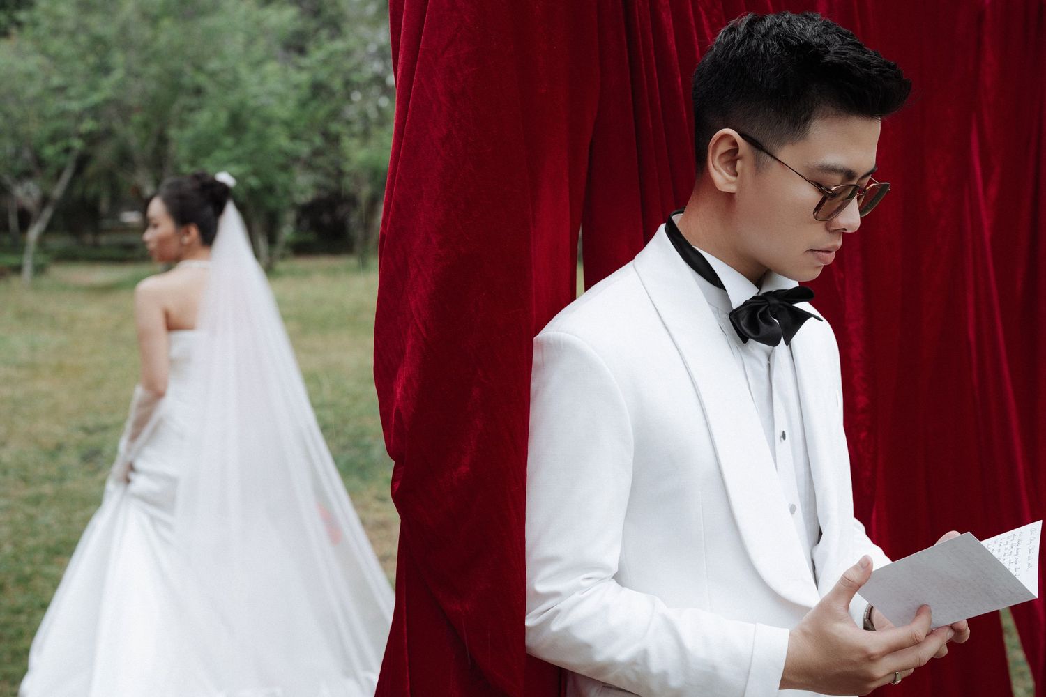Groom in white tuxedo reads vows from a card while bride stands before red draped backdrop outdoors.