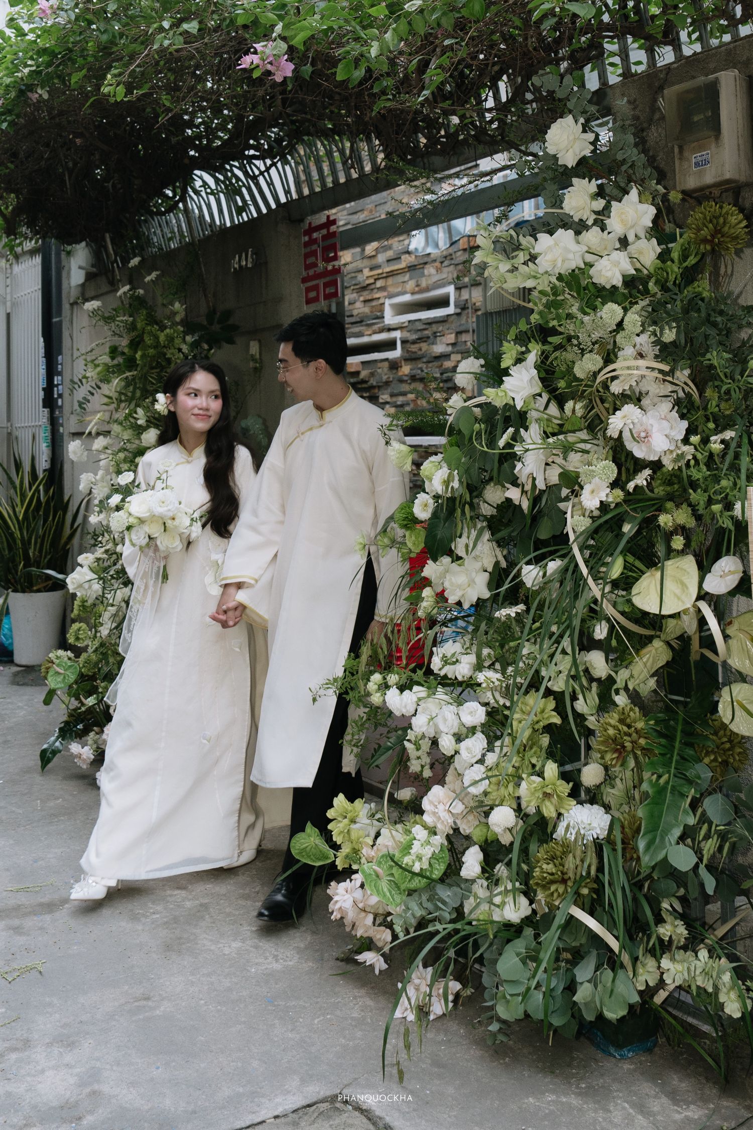 Vietnamese bride and groom in white ao dai walk hand in hand past stunning white floral arch display.