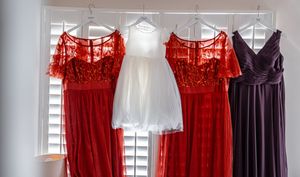 Several formal dresses in red, white, and navy blue hang from hangers against a white wall near venetian blinds.