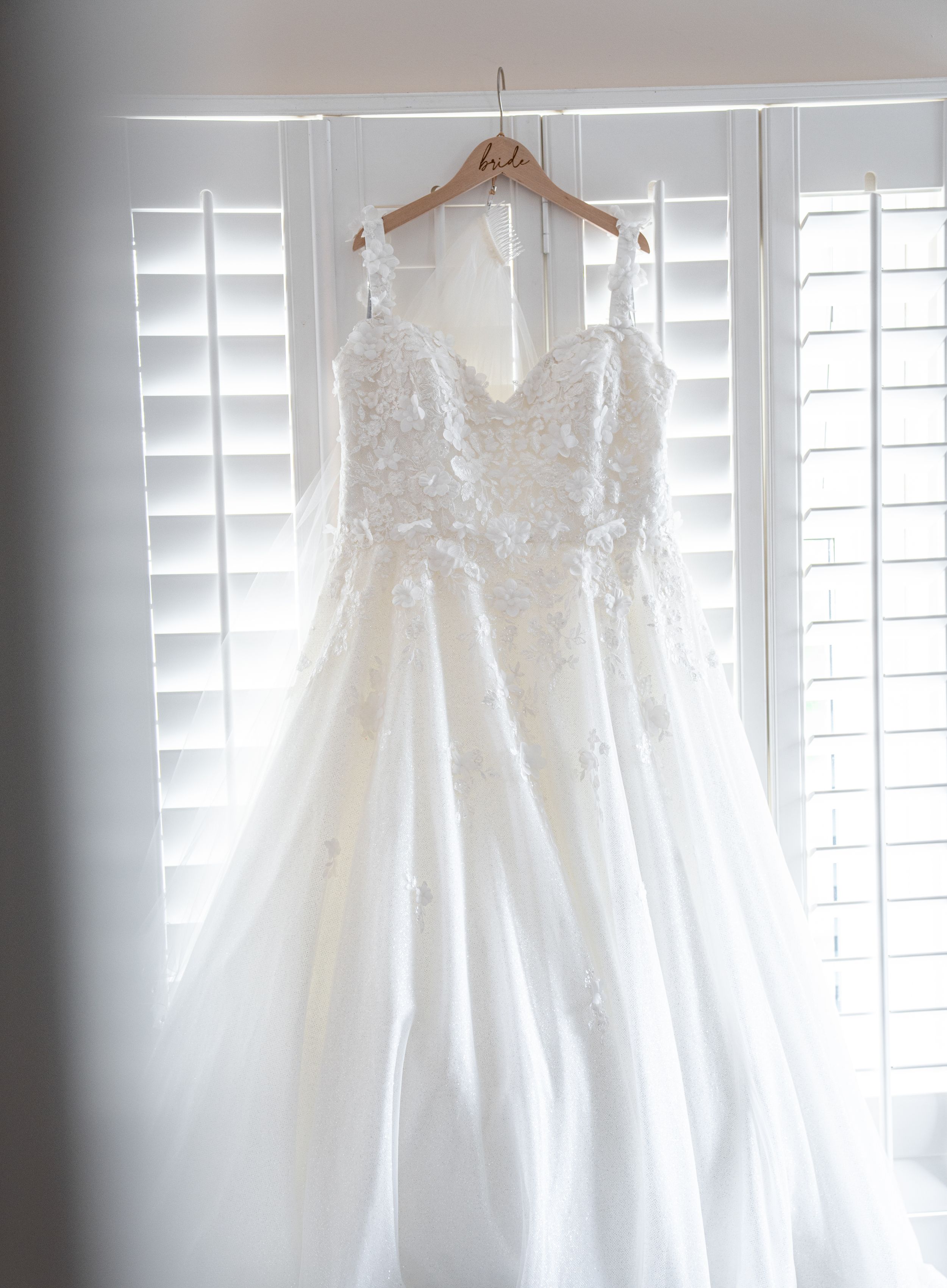 Five elegant white lace wedding dresses hanging on wooden hangers against a bright window with white shutters.