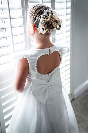 Back view of a white flower girl dress with delicate bow detail and elegant updo hairstyle near window.