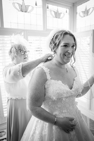 Black and white image showing a bride in a wedding dress during preparation with natural light streaming through windows.