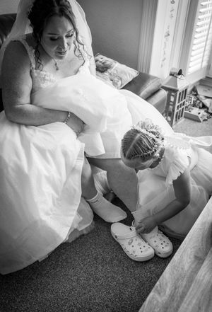 Black and white photo of wedding preparation showing white Crocs being put on with dress in frame.