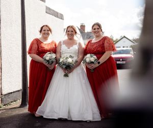 A row of bridesmaids in burgundy lace dresses stand with a bride in a white gown holding bouquets at a wedding.